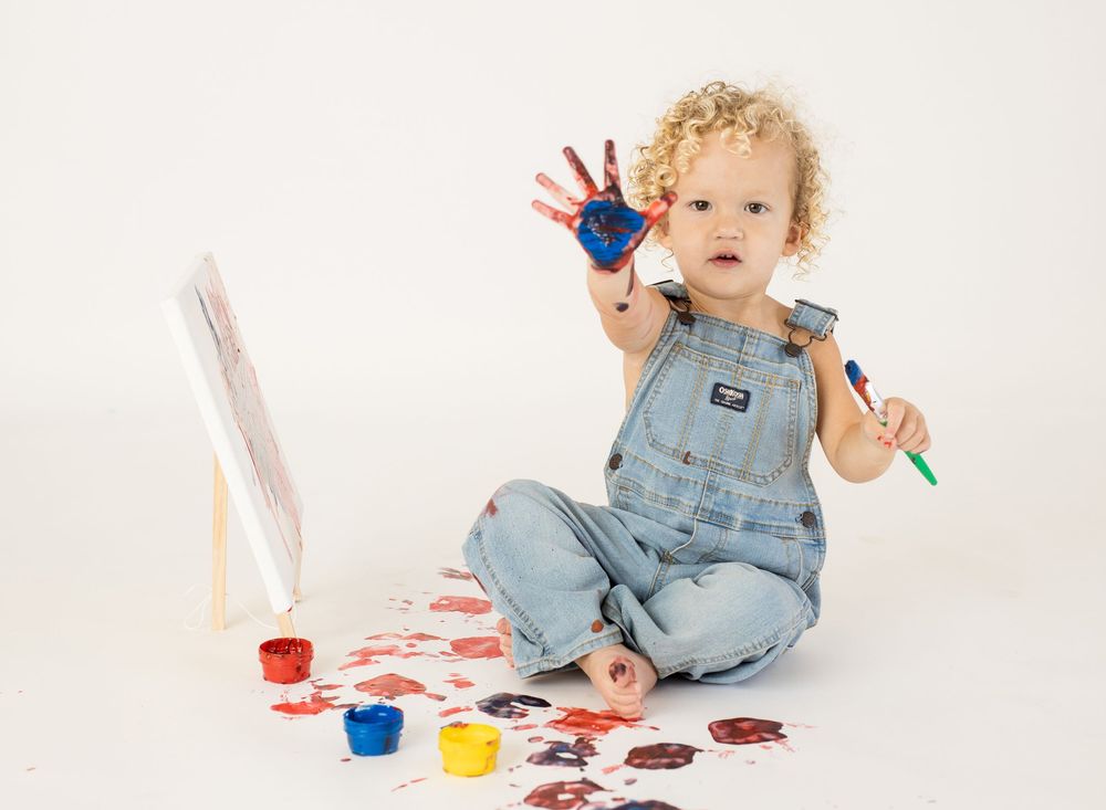 Toddler in denim overalls with paint-covered hands sitting by an easel with colorful paint jars.