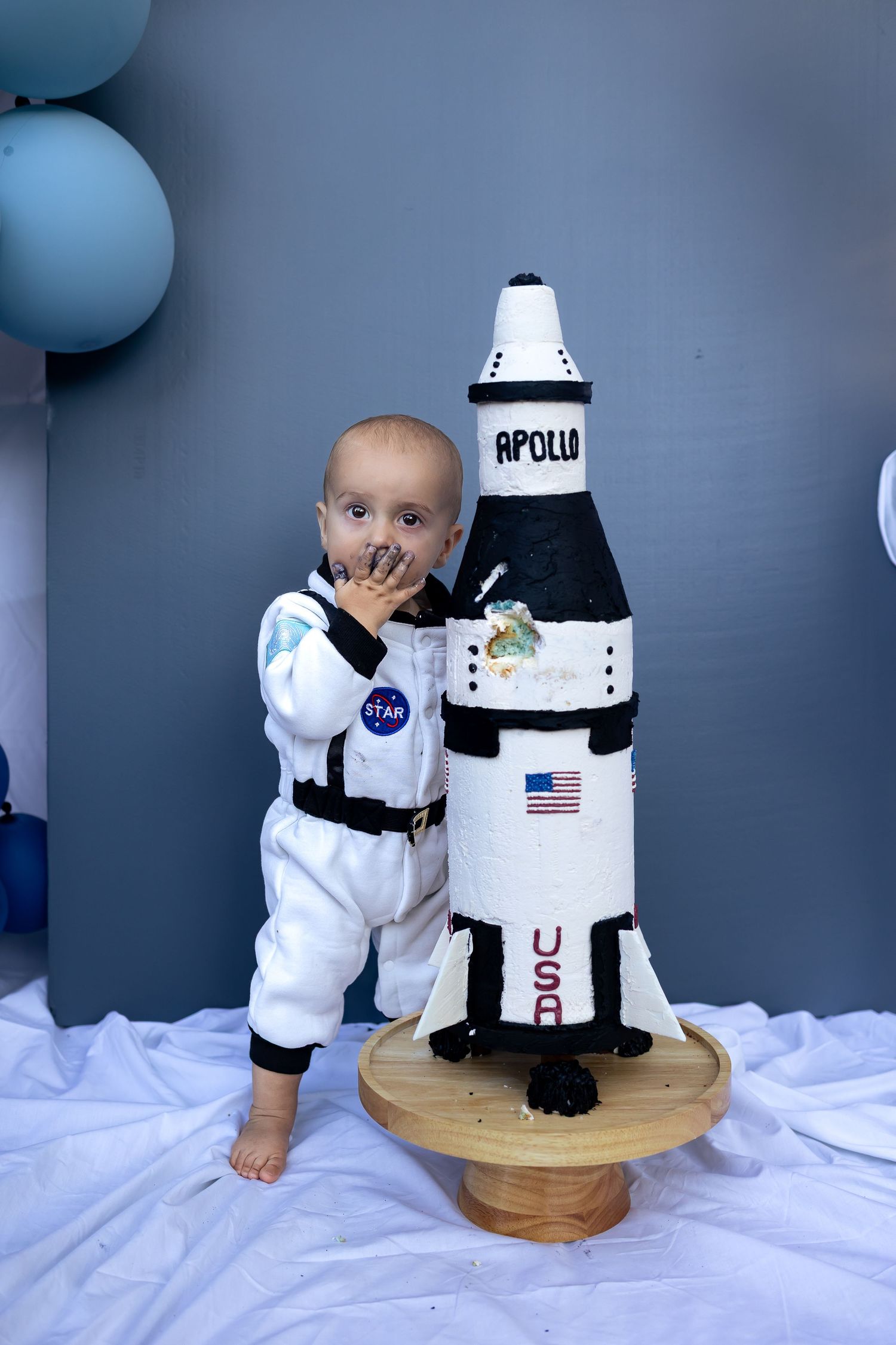 Baby in NASA astronaut outfit stands beside an Apollo rocket cake topper at a space-themed first birthday party.