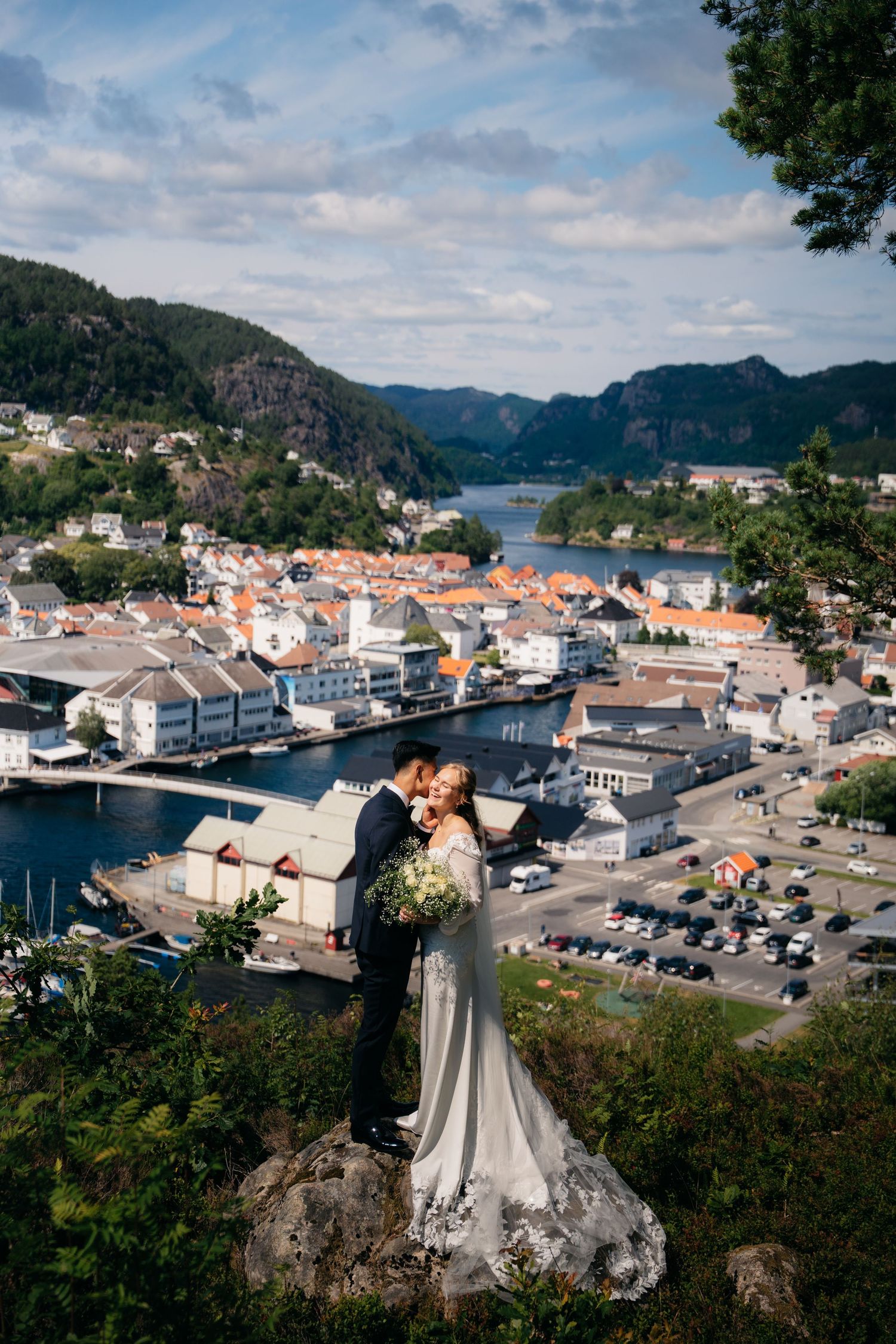 Bride and groom embrace on a rocky overlook with a stunning Norwegian fjord town backdrop on their wedding day.