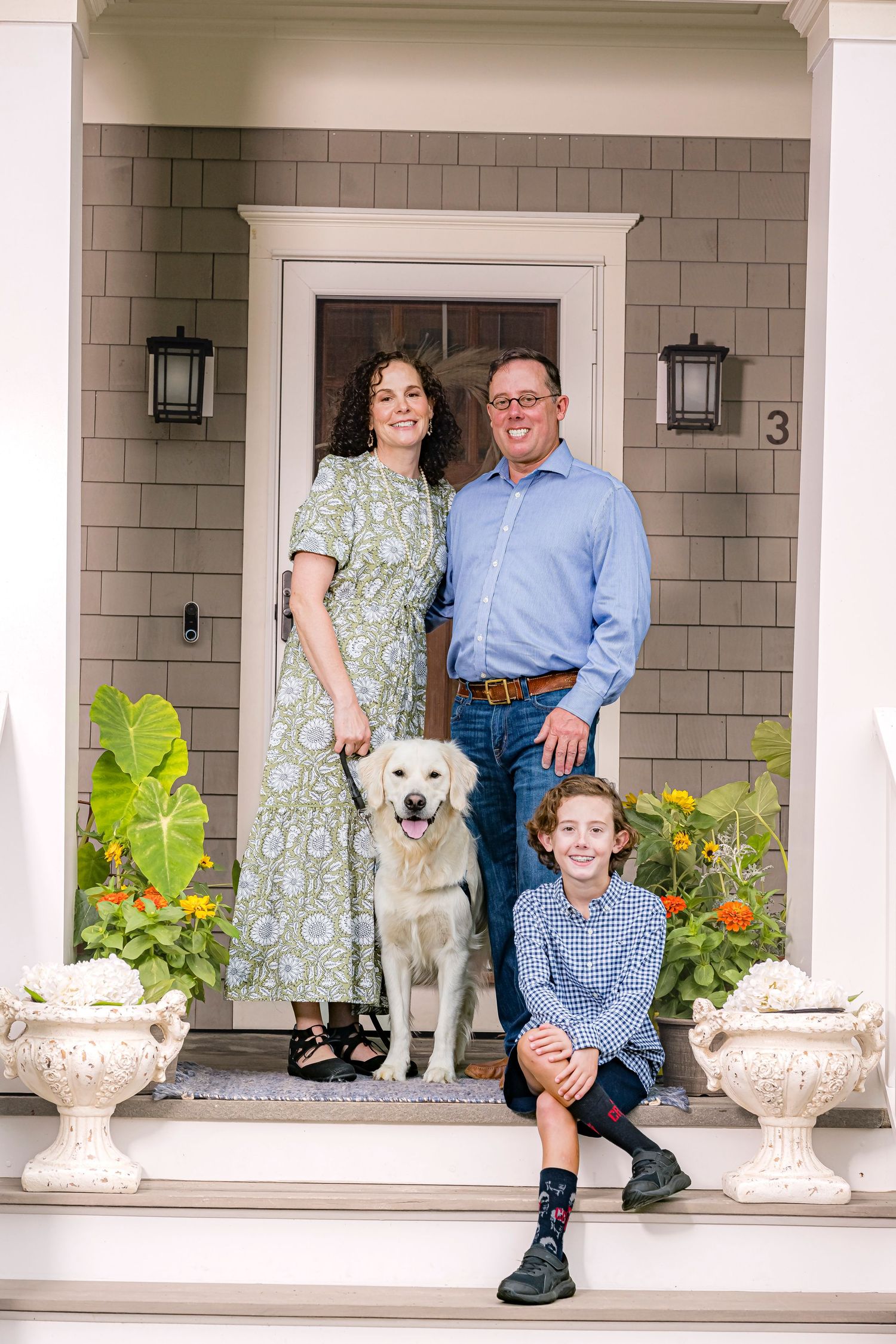 Family of three smiling on a white porch railing outside a gray house with flowers.