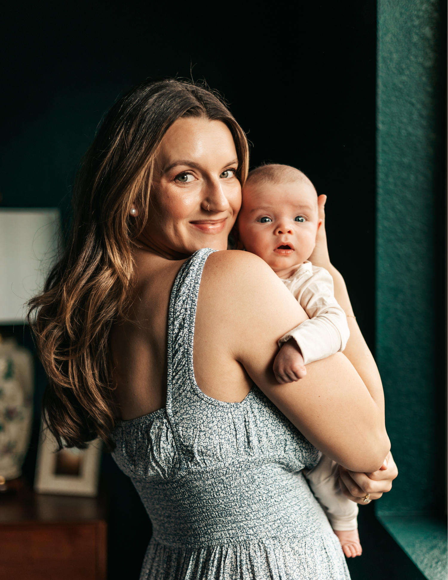 A smiling brunette woman in a gray dress holds a newborn baby over her shoulder indoors.