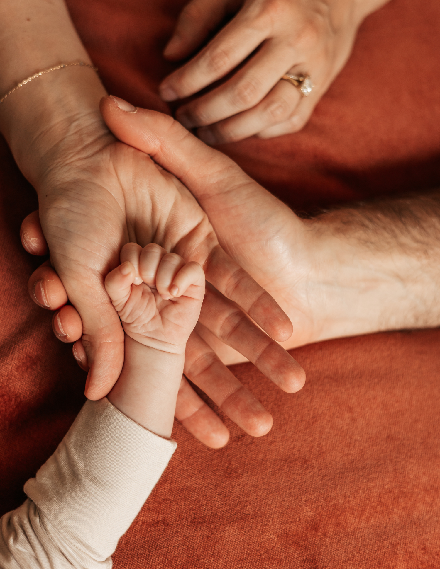 A newborn baby's tiny hand resting in the open palms of two parents wearing wedding rings on a red background.