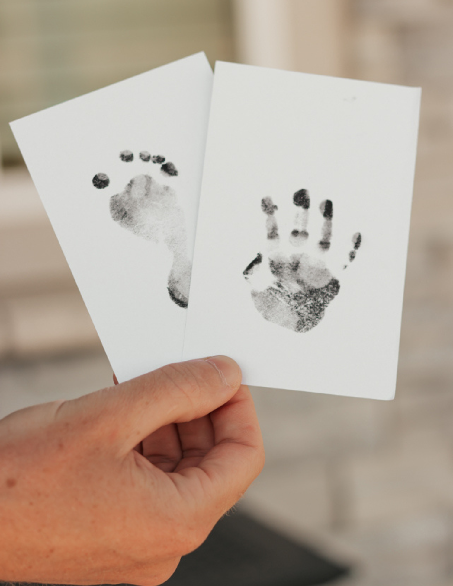 Hand holding two white cards with black ink baby footprint and handprint stamps on them.