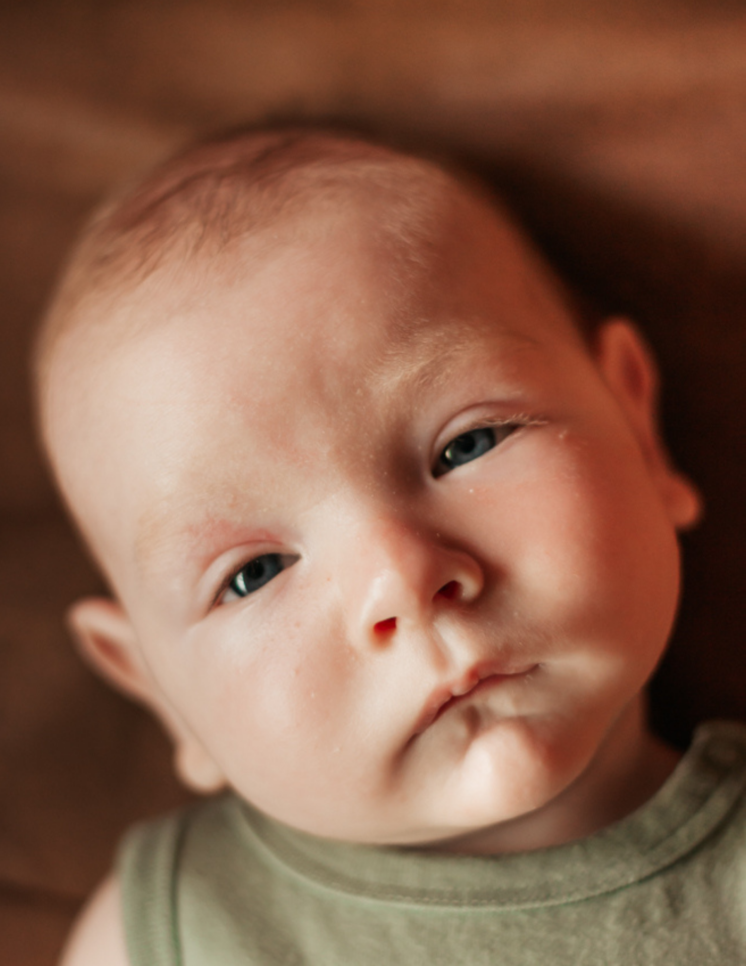 Close-up portrait of a curious newborn baby with blue eyes wearing a green onesie, lying on a warm wooden surface.