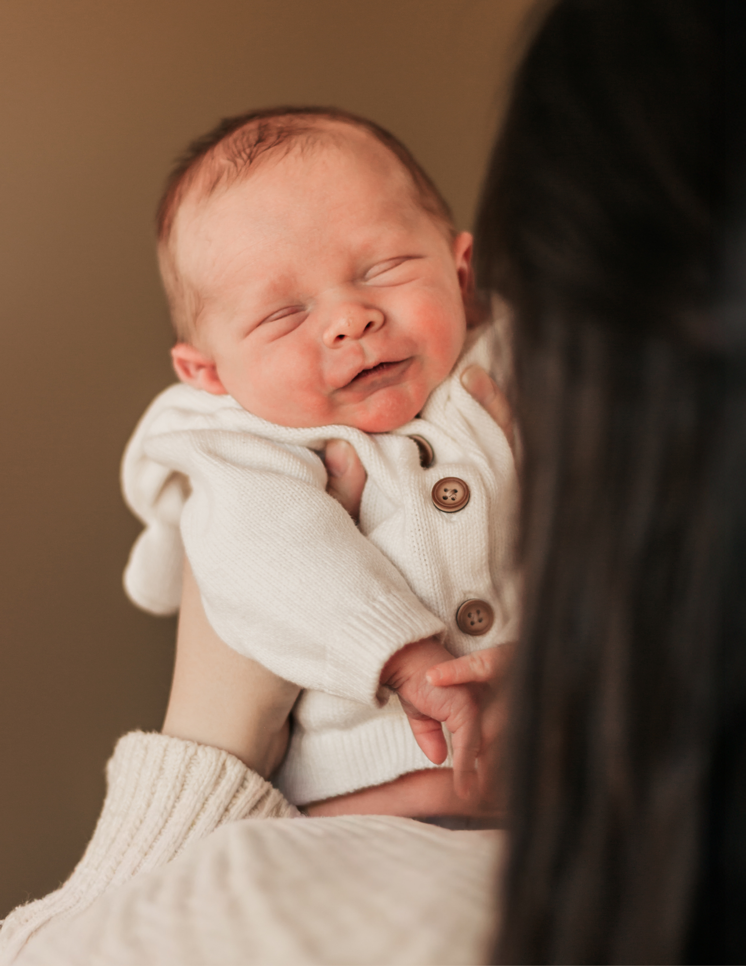 A newborn baby in a white knit outfit smiles while being held up by a dark-haired mother in a warm studio.