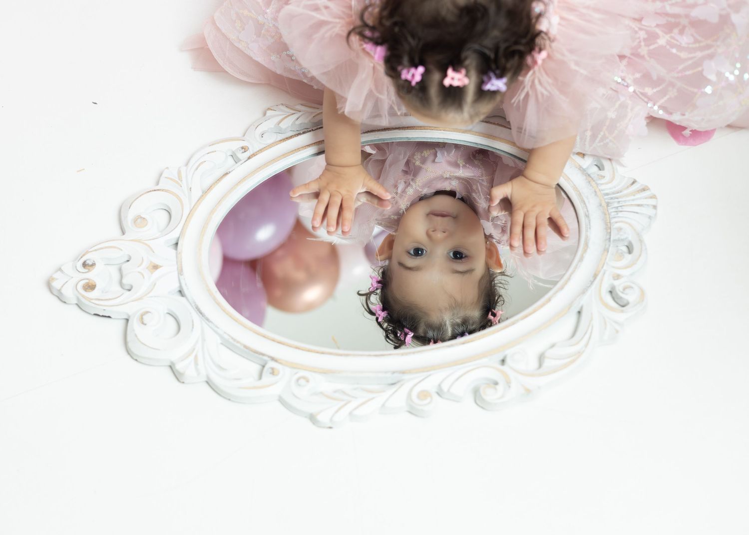 Toddler girl in pink dress reflected in ornate white mirror with balloons during first birthday photo shoot.
