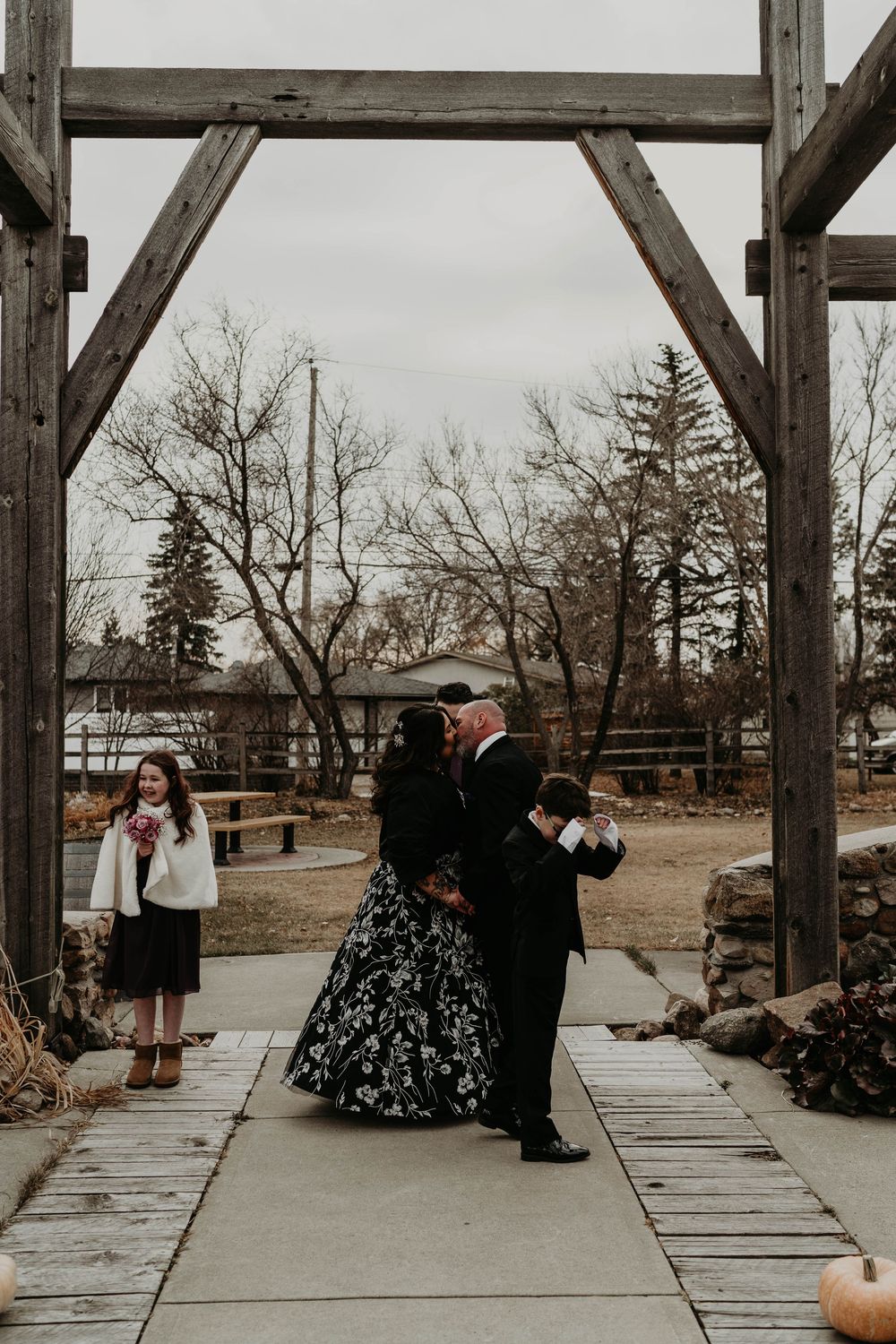 Couple sharing first kiss in black floral wedding attire under rustic wooden arch at outdoor fall ceremony.