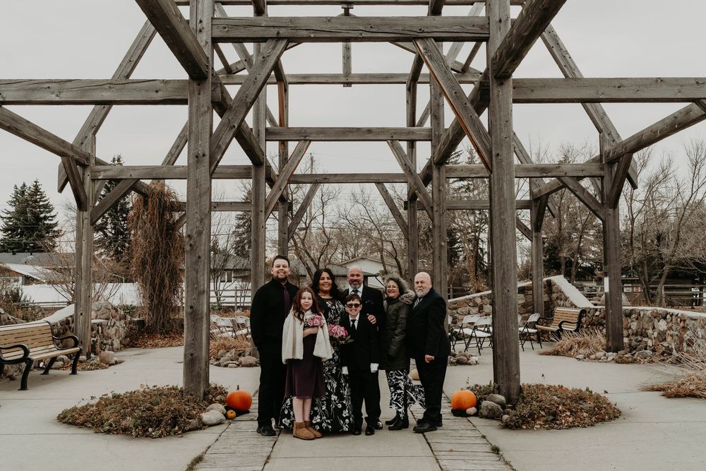 Family group portrait under rustic wooden pergola surrounded by fall pumpkins at outdoor wedding venue.