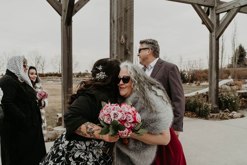 Two women embrace at an outdoor wedding ceremony, one holding pink roses, while guests look on under a wooden pergola.