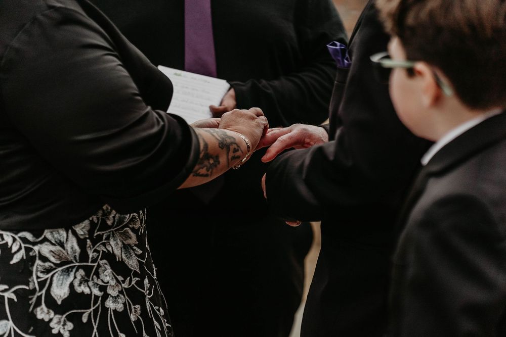 A tattooed officiant places a wedding ring on a bride's hand during a ceremony, with a young boy in a suit watching.