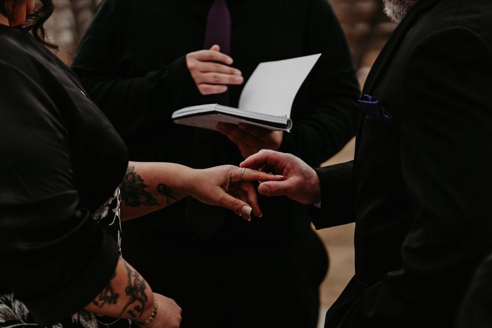 Close-up of a wedding ring exchange ceremony, with tattooed hands and officiant holding an open book.