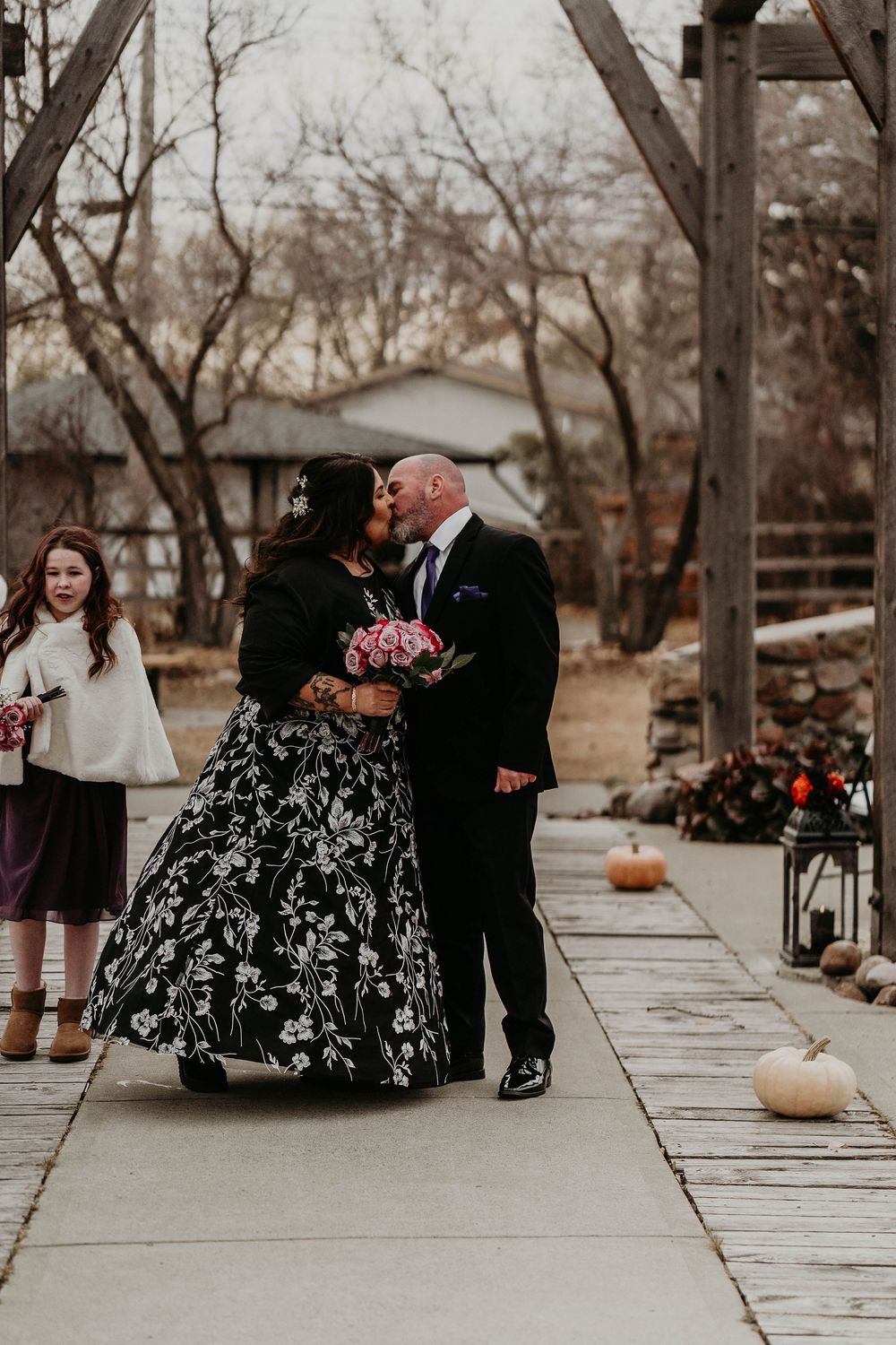 Couple sharing first kiss in black floral wedding dress and suit on outdoor ceremony path with pumpkins.