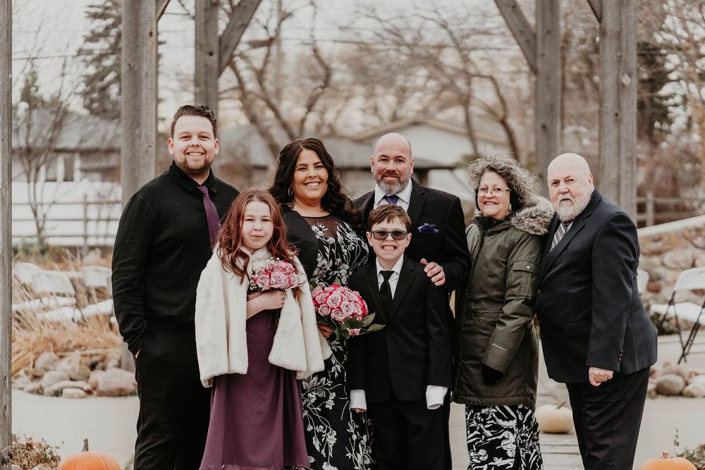 Family group portrait at an outdoor wedding venue in autumn, posing under a pergola with fall foliage.