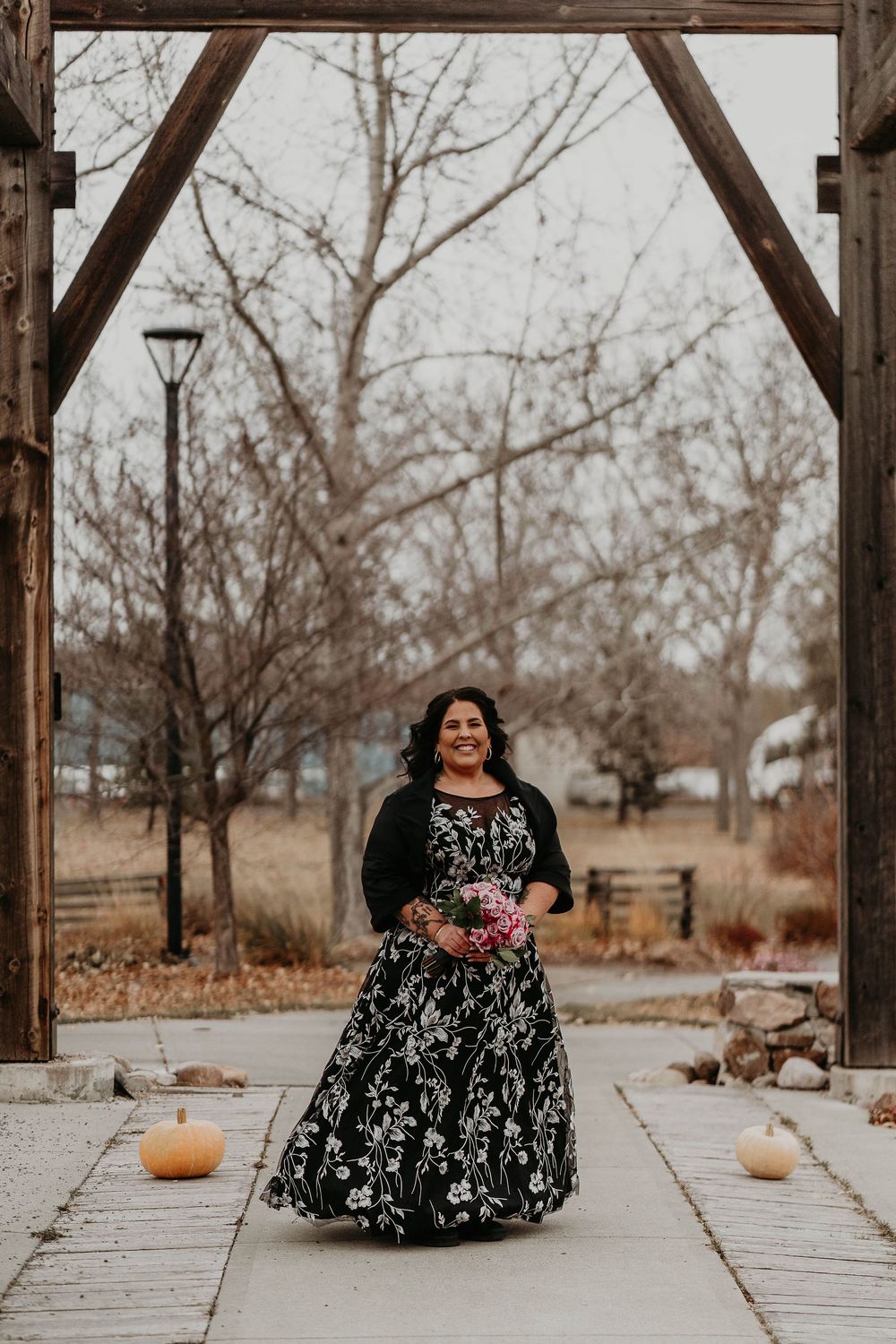 Woman in black floral gown holding bouquet walks under rustic wooden arch at outdoor autumn wedding venue.