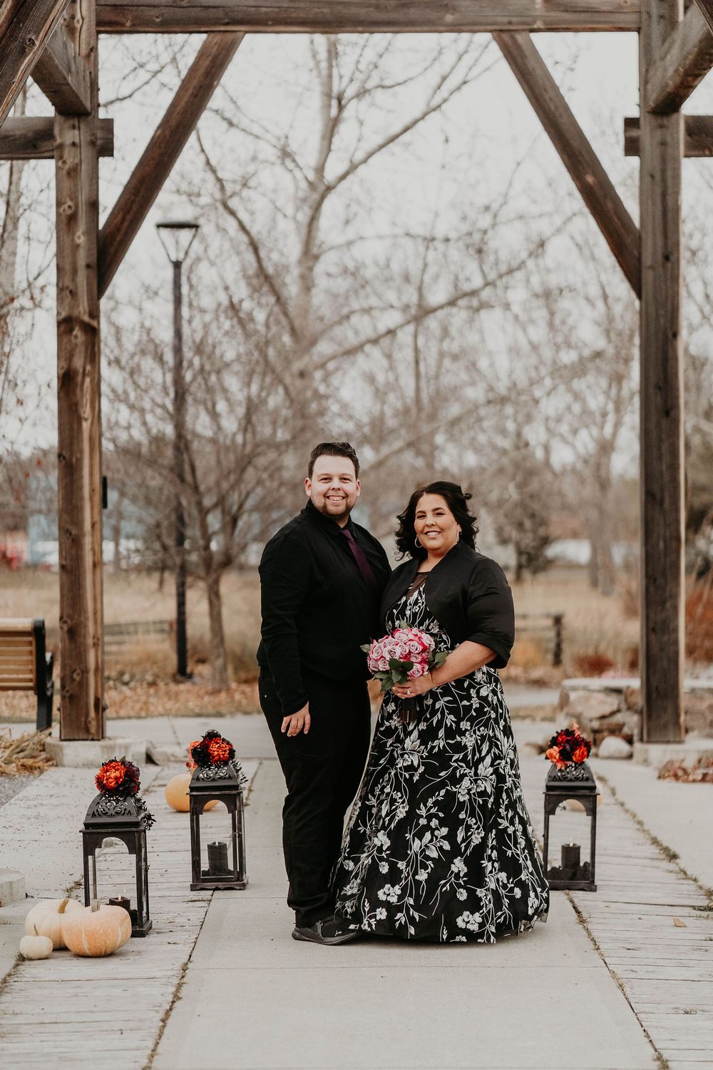 Couple posing under rustic wooden arch with lanterns and pumpkins, woman in floral gown holding bouquet, man in black suit.