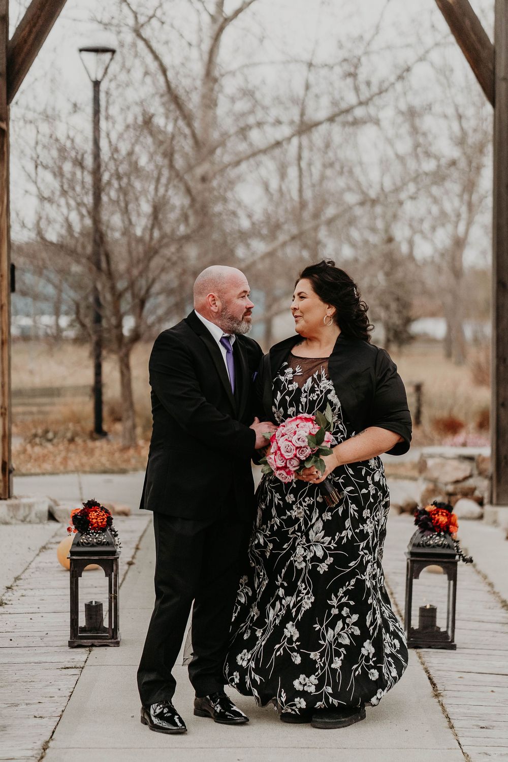 Couple in formal attire smiling at each other outdoors, woman holding pink bouquet in black floral dress, man in black suit.