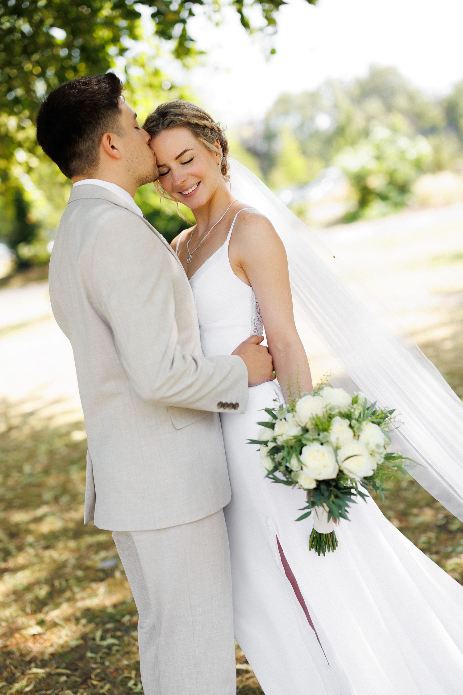 Happy newlyweds walk outdoors, bride in white gown holding bouquet, groom in beige suit, smiling in sunny park.