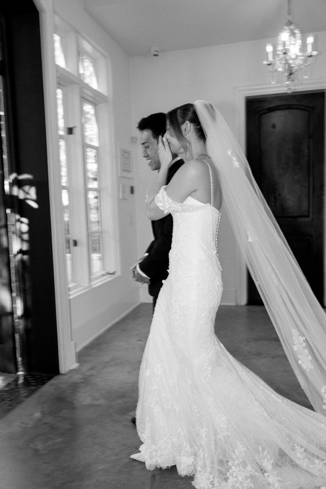 Black and white photo of a bride in a lace gown and veil standing with a groom in a tuxedo indoors.