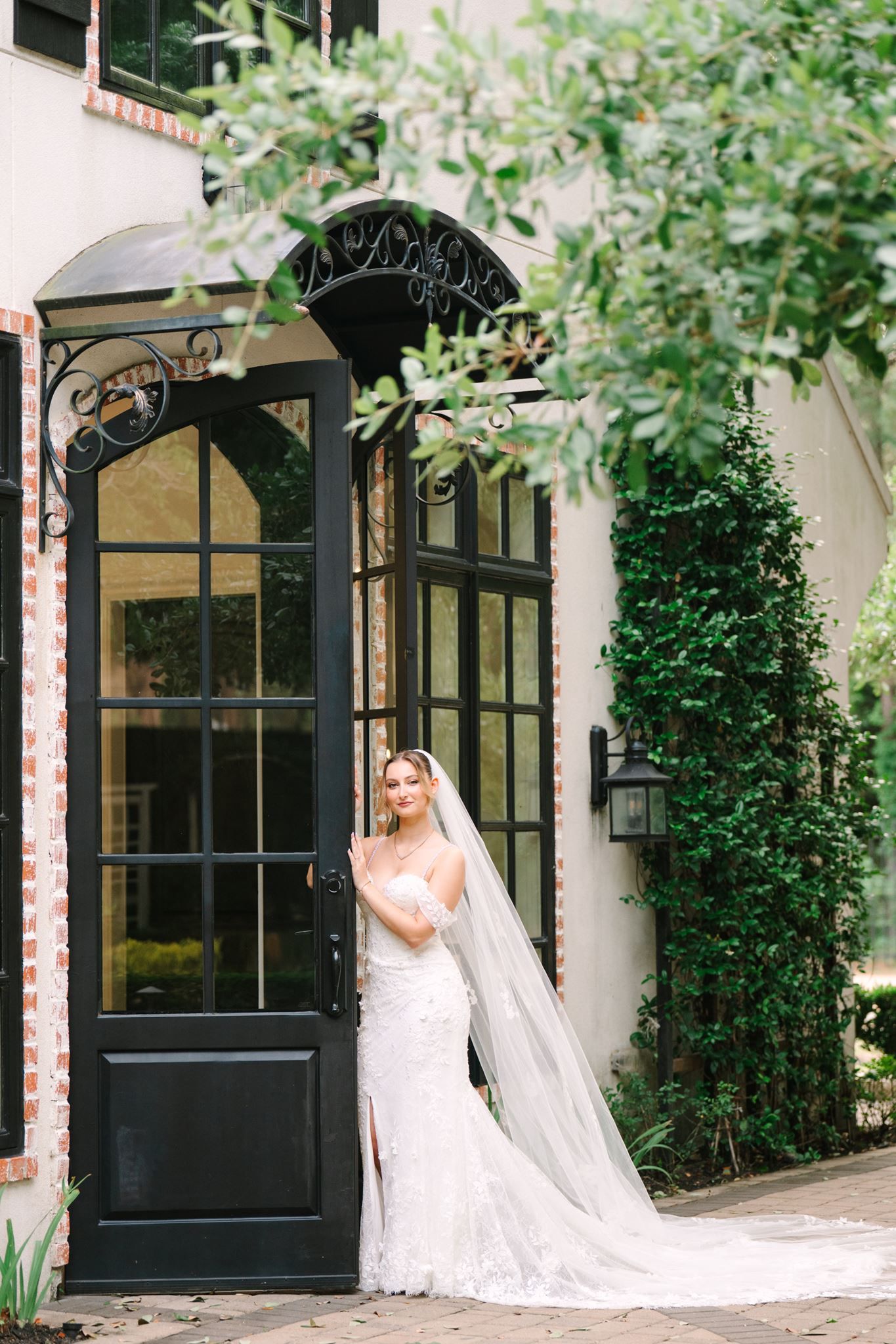 Bride in lace mermaid gown and cathedral veil poses by elegant black French door with ivy-covered wall.