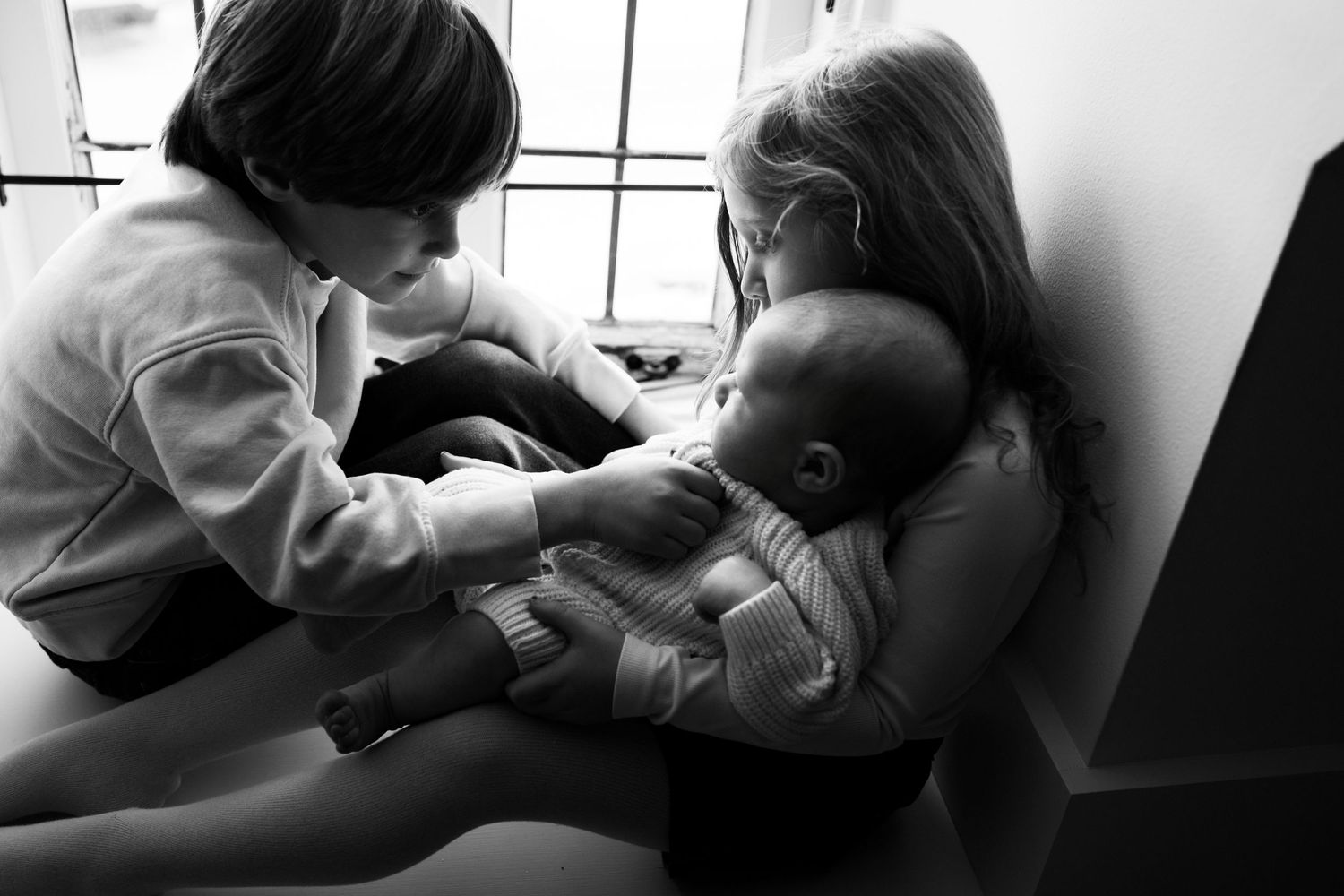 Black and white photo of two children tenderly holding a newborn baby indoors.