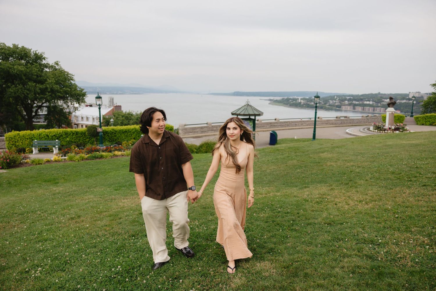 Couple holding hands walking on green grass with scenic waterfront and cloudy sky in background.