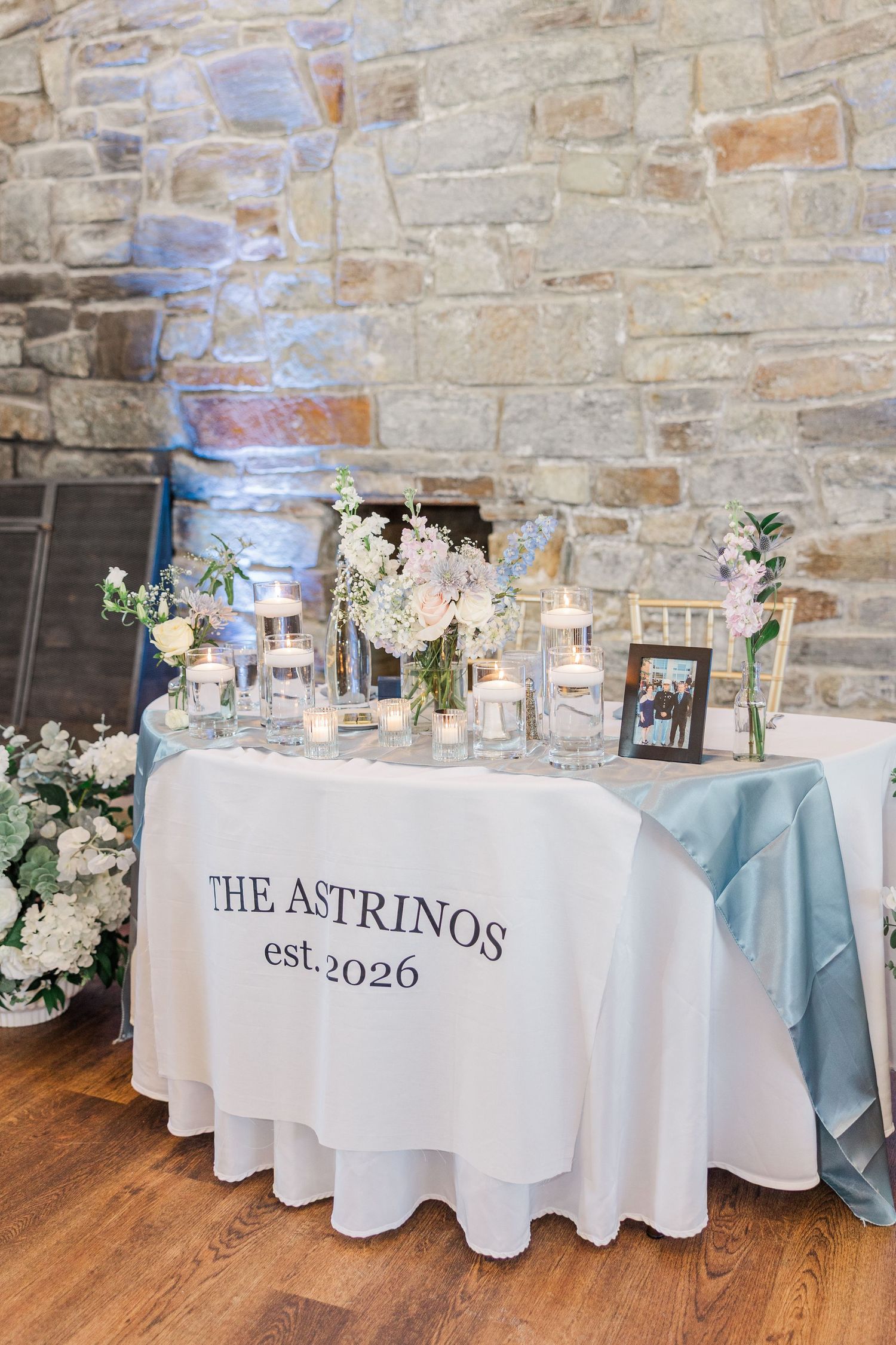 Elegant wedding sweetheart table with white linen, 'The Astrinos est. 2026' text, candles, and floral arrangements.