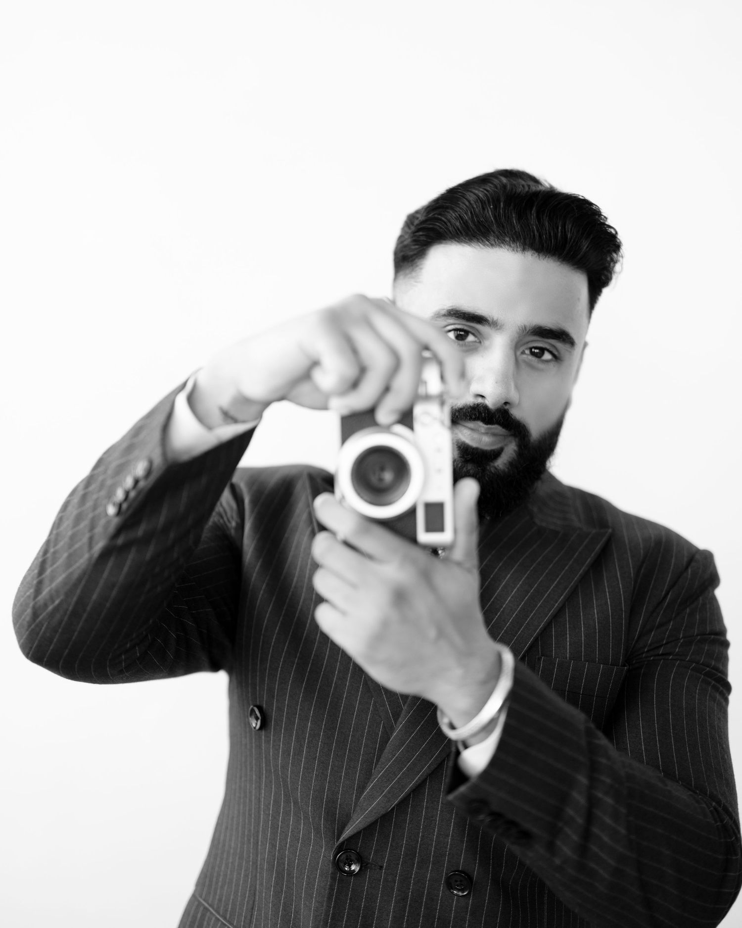 Black and white portrait of a bearded man in a pinstripe suit holding a vintage film camera up to take a photo.