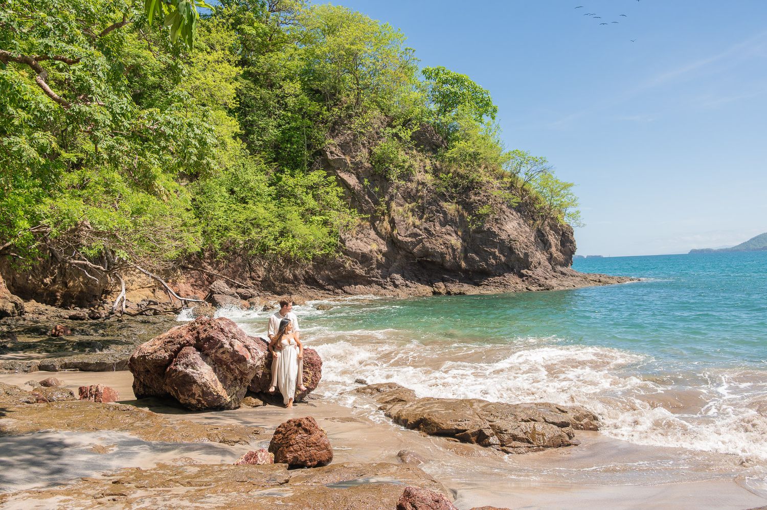 Couple in wedding attire standing on rocky tropical beach with lush green cliffs and turquoise ocean waves.