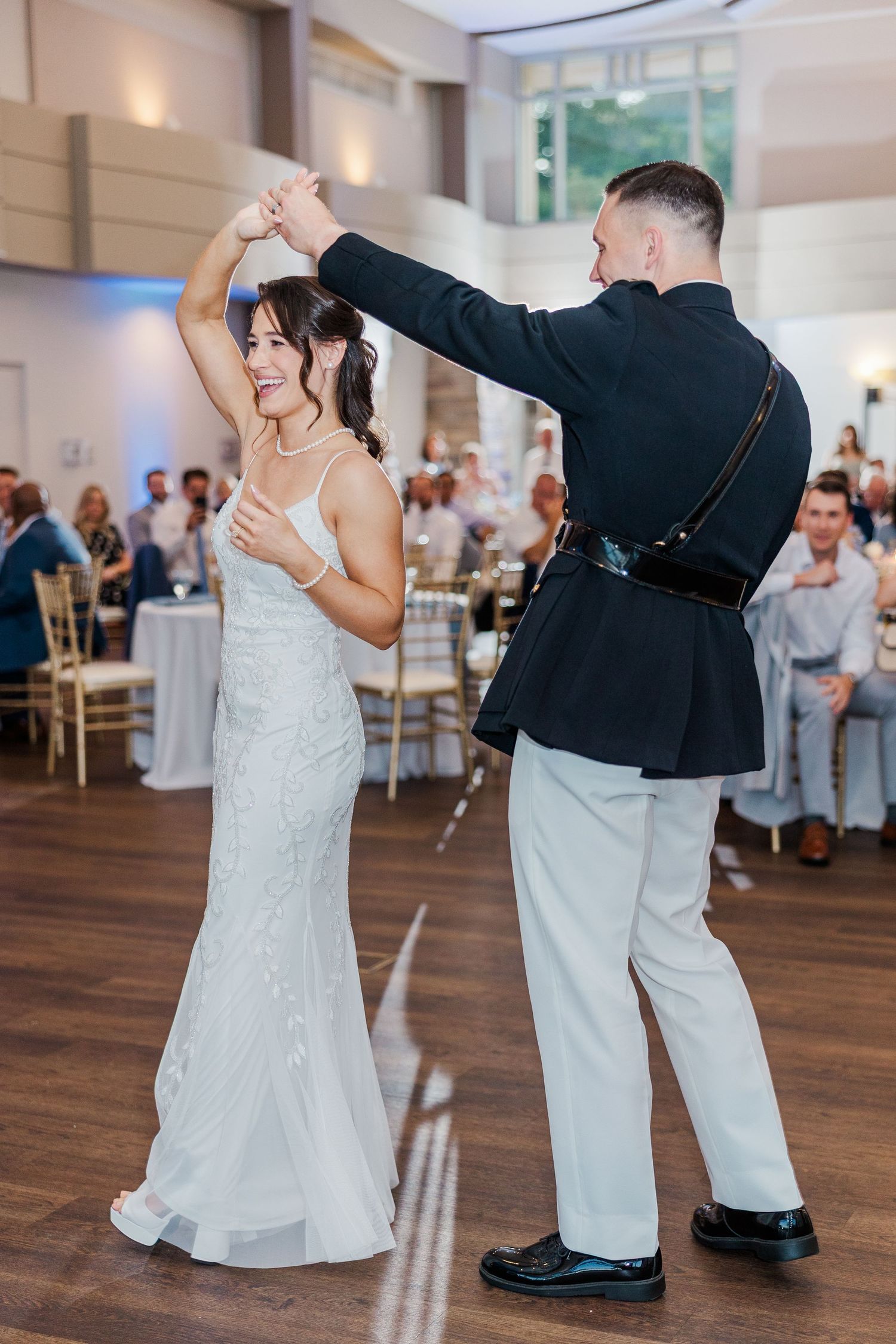 Bride and groom share first dance at wedding reception, groom in military dress uniform twirling bride in white gown.