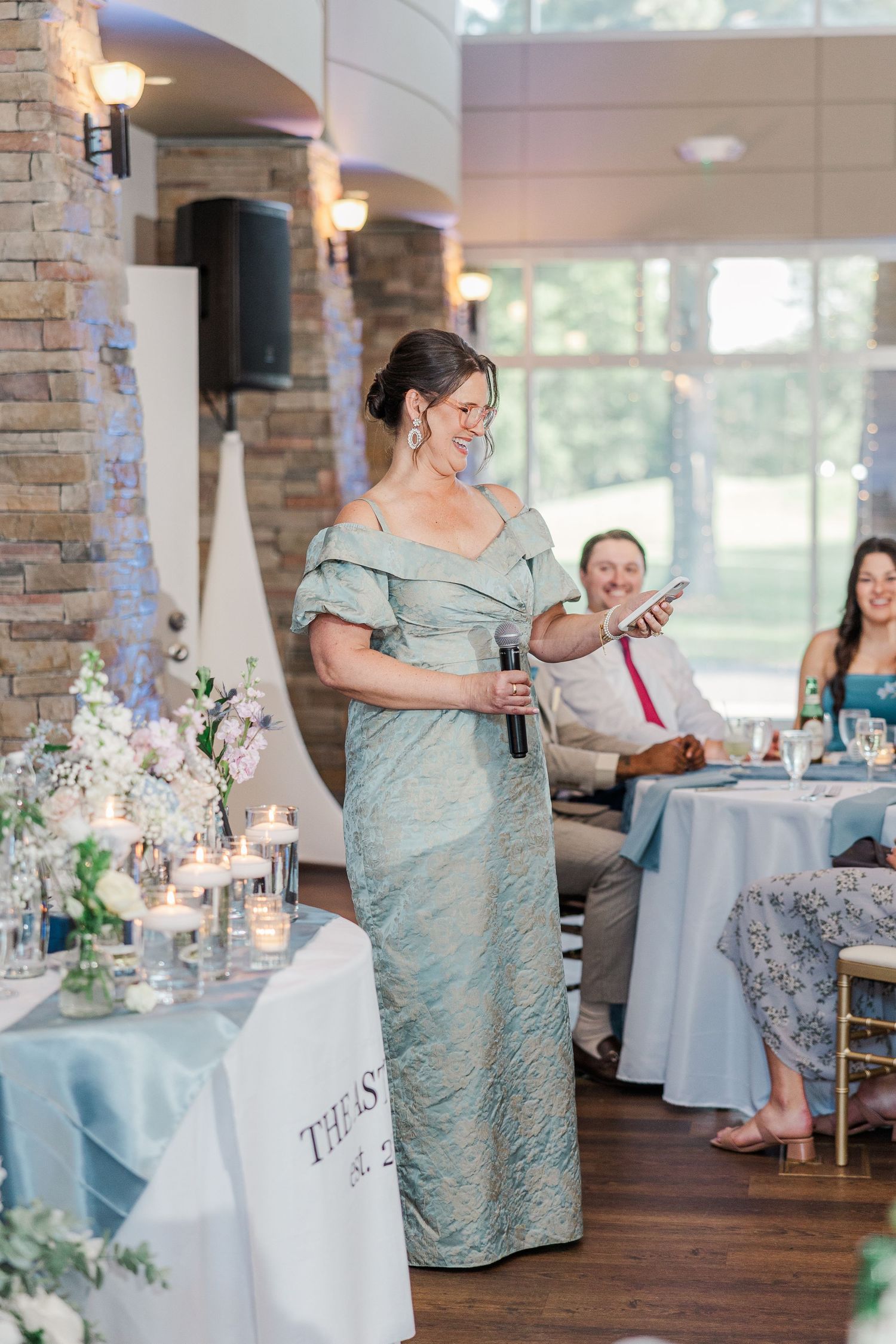 Woman in sage green gown giving a speech at an elegant wedding reception with candlelit tables and floral arrangements.