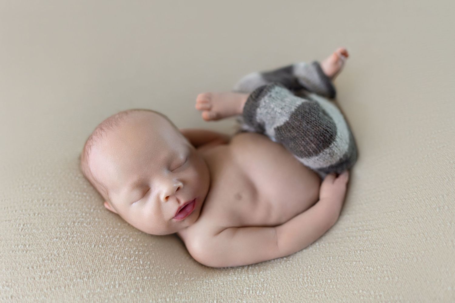 Sleeping newborn baby wearing gray knit pants posed on a beige blanket in a professional newborn photography session.