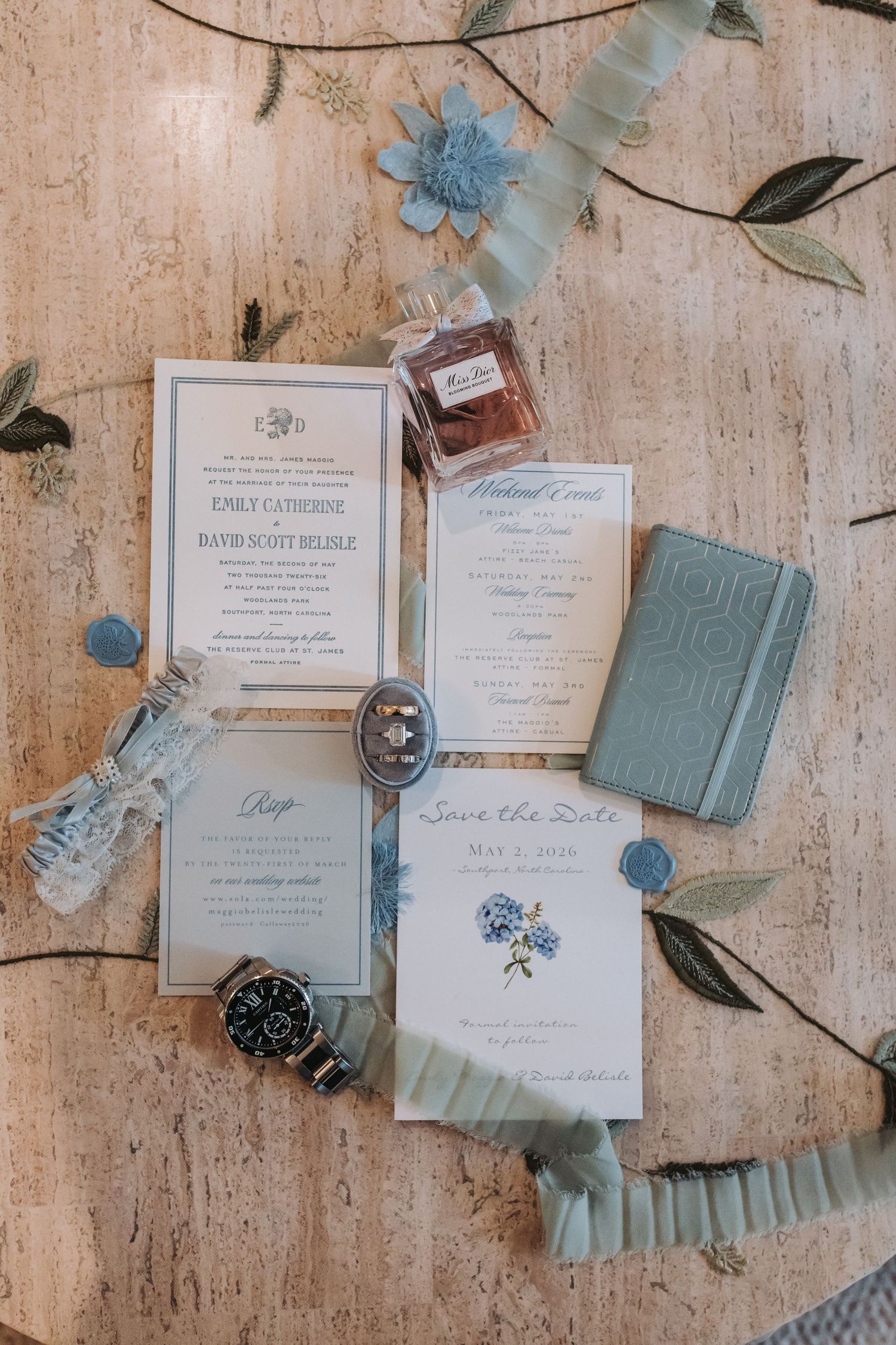 Elegant wedding stationery flat lay featuring blue hydrangea save the date cards, invitations, perfume, watch, and ribbon.