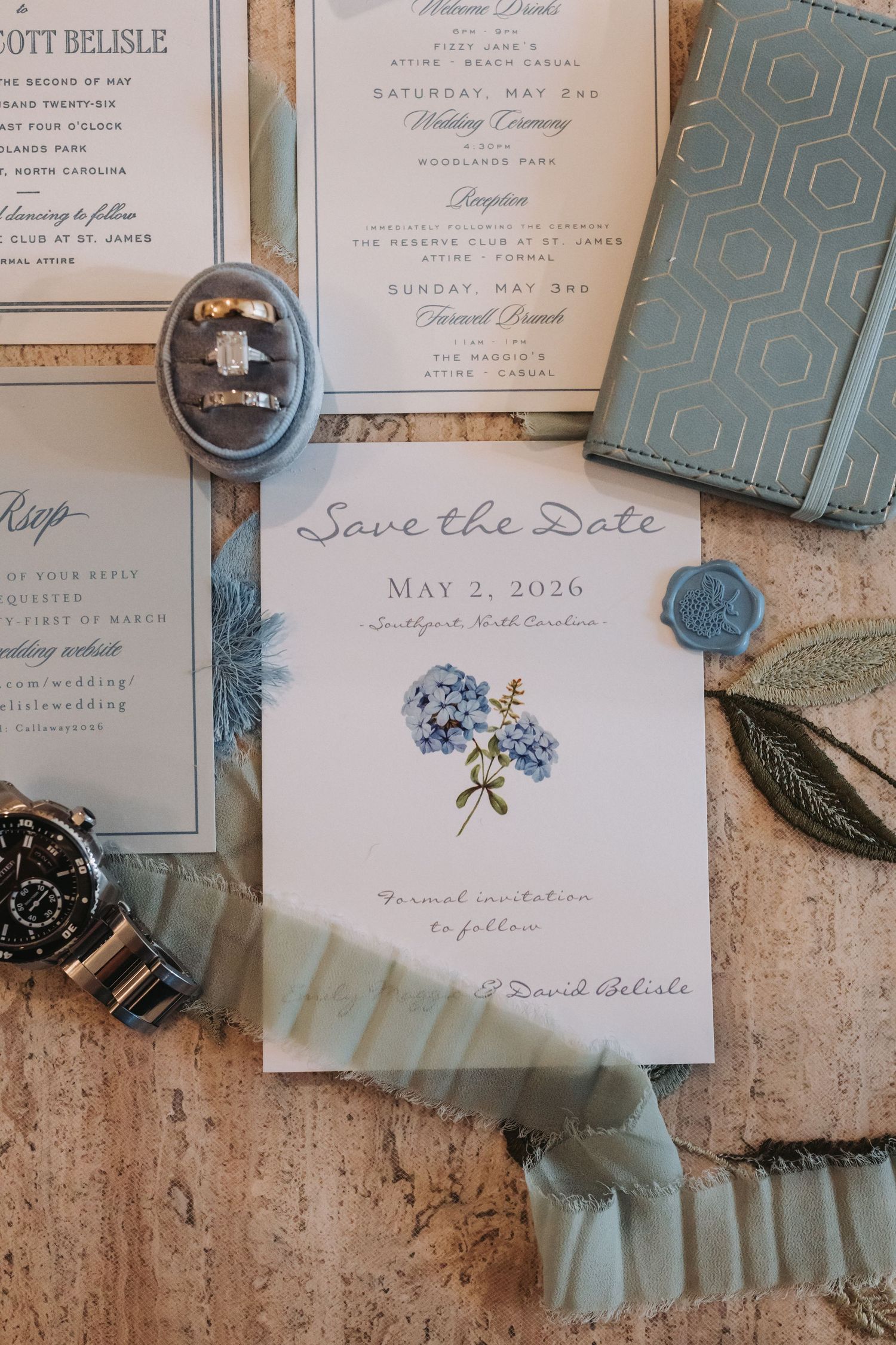 Elegant wedding save the date card for May 2, 2026 with blue hydrangea illustration, surrounded by rings, watch, and stationery.