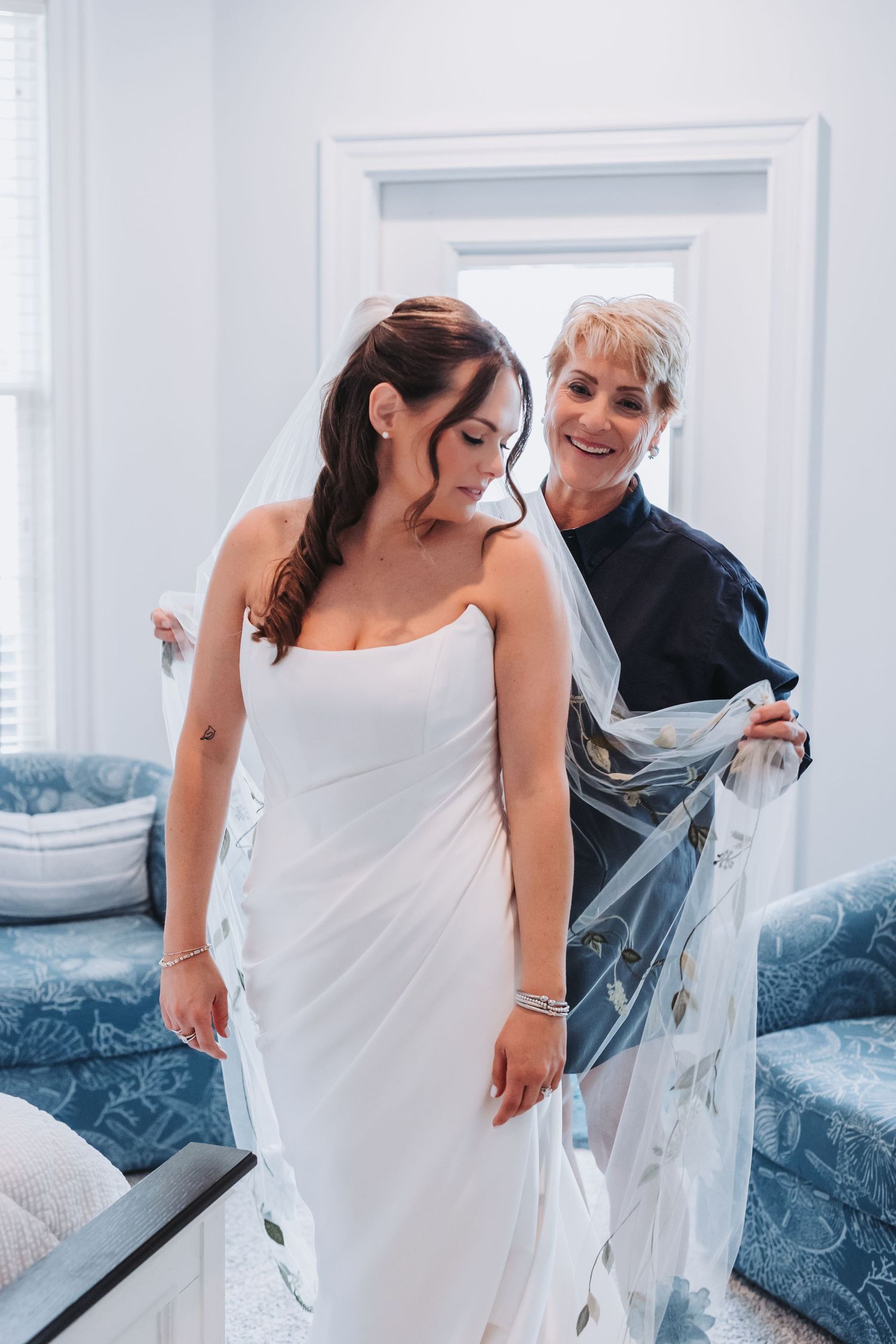 Smiling older woman in navy dress holds floral veil behind brunette bride in white strapless wedding gown.
