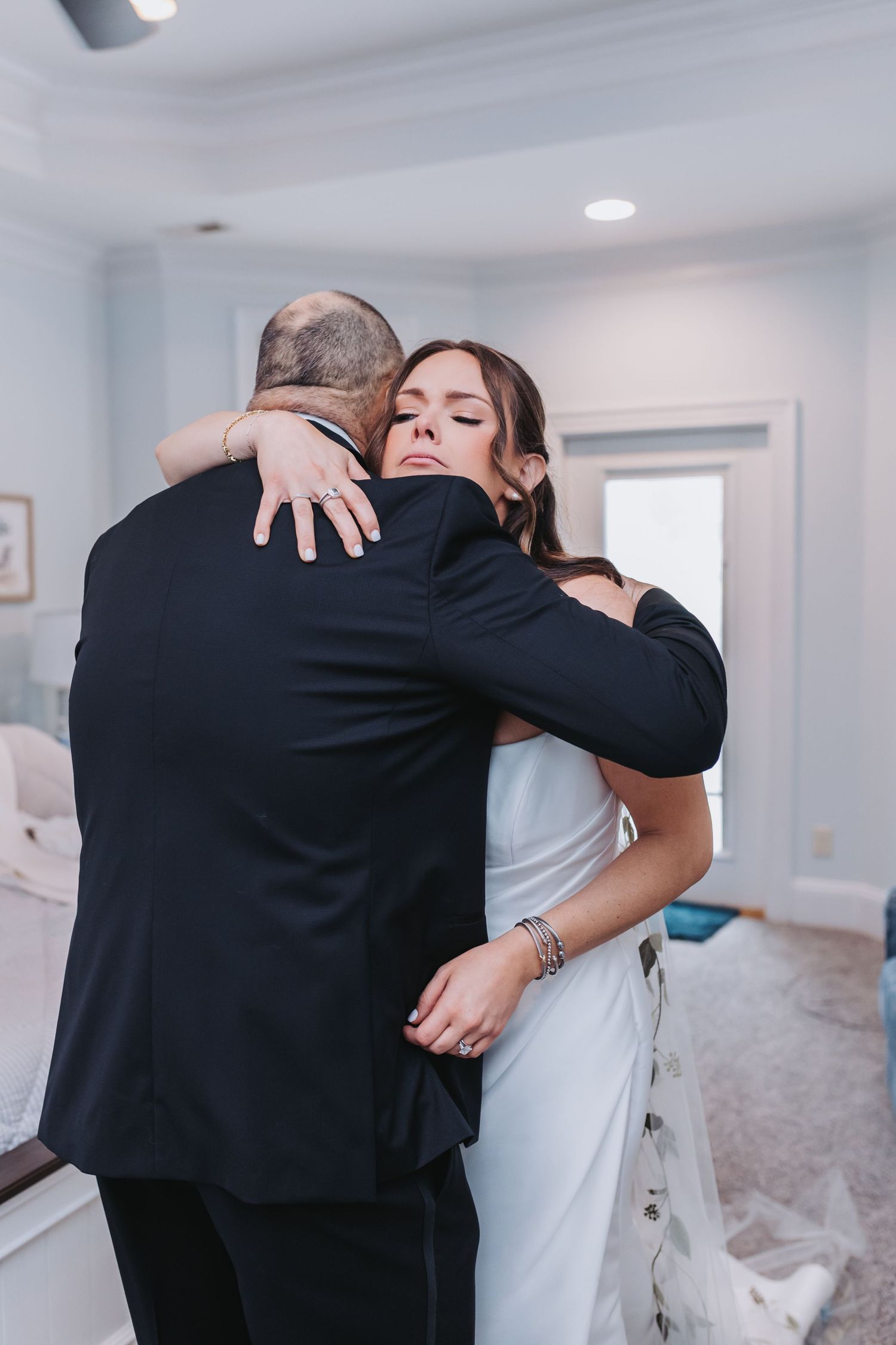 Emotional bride in white gown embraces man in black tuxedo during heartfelt first look moment indoors.