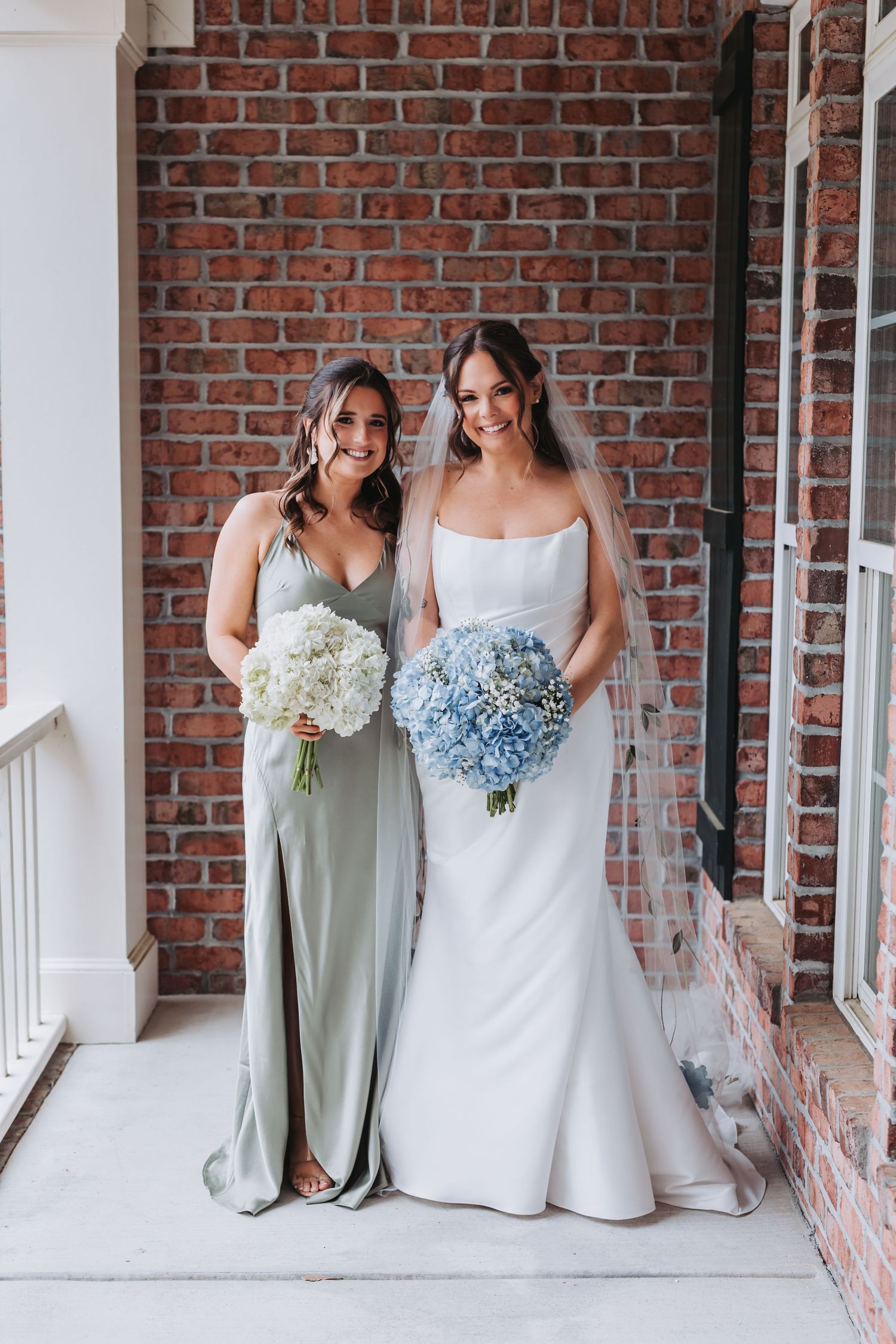 Bride in white strapless gown holding blue hydrangea bouquet poses with bridesmaid in sage dress against brick wall.