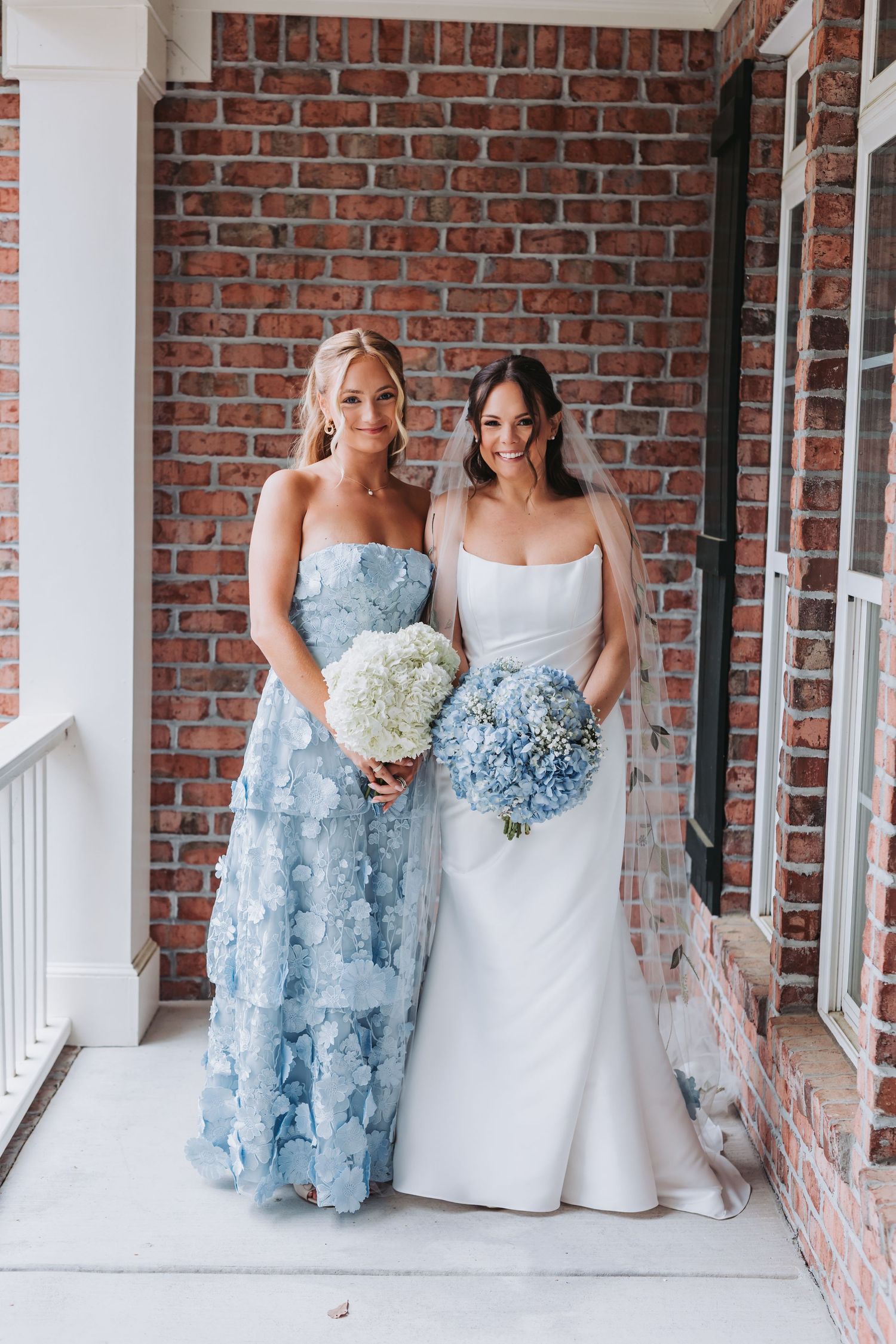 Bride in white gown and bridesmaid in blue floral dress holding bouquets on brick building porch.