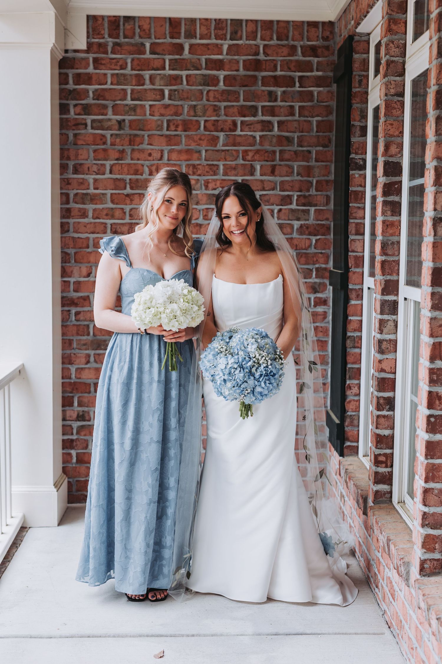Bride in white gown and bridesmaid in blue dress holding hydrangea bouquets against brick wall.