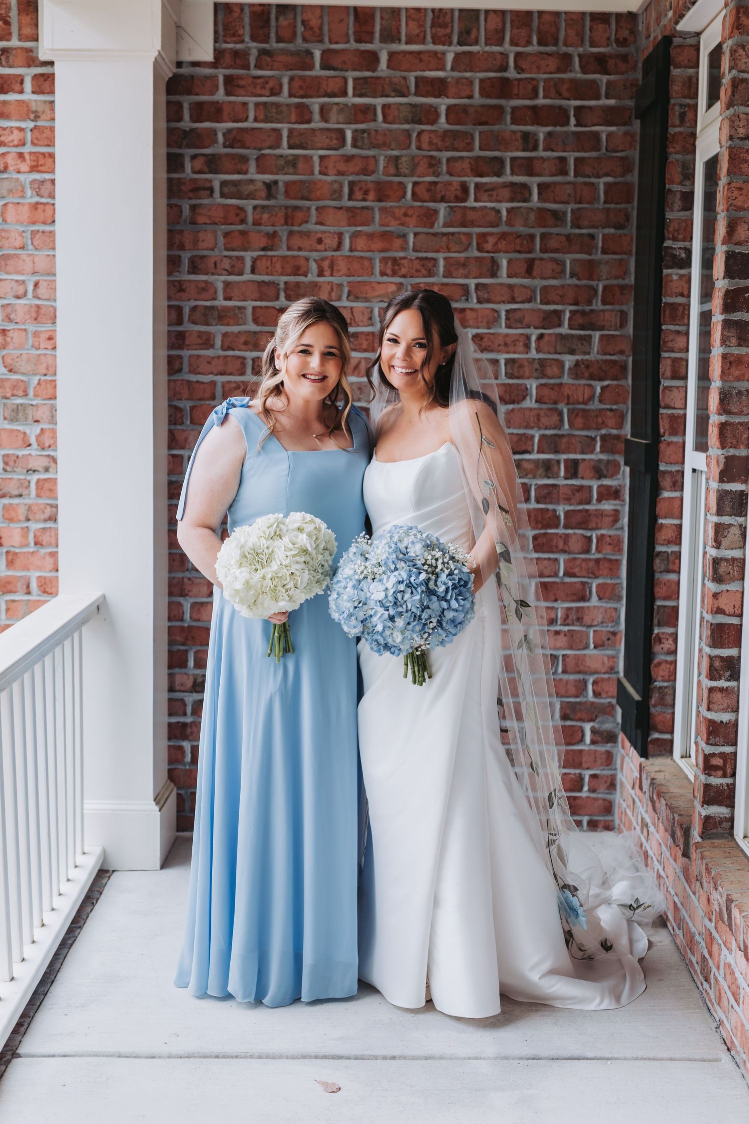 Smiling bride in white gown and bridesmaid in light blue dress holding hydrangea bouquets on a brick porch.
