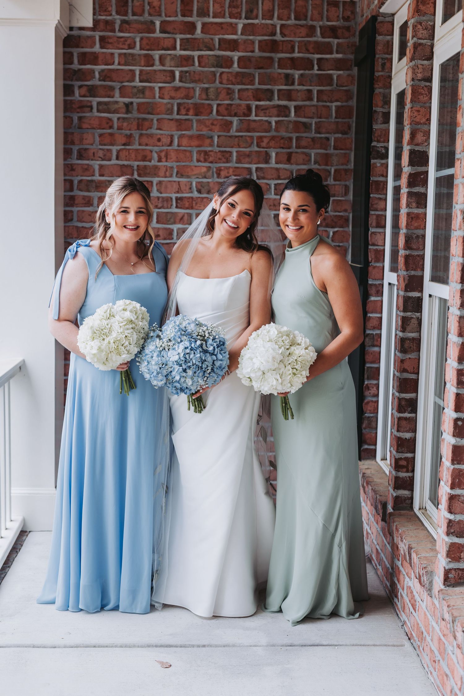 Bride with two bridesmaids in blue and sage green gowns holding white hydrangea bouquets outdoors.