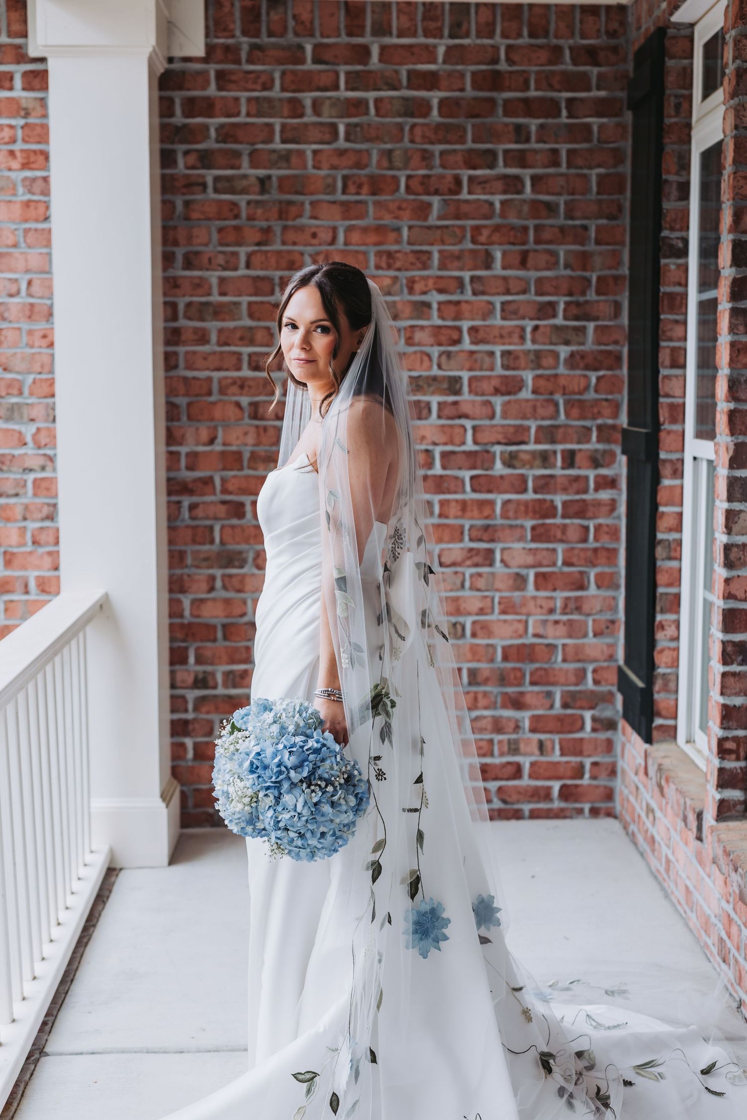 Bride in white gown with floral veil holding blue hydrangea bouquet against brick wall.
