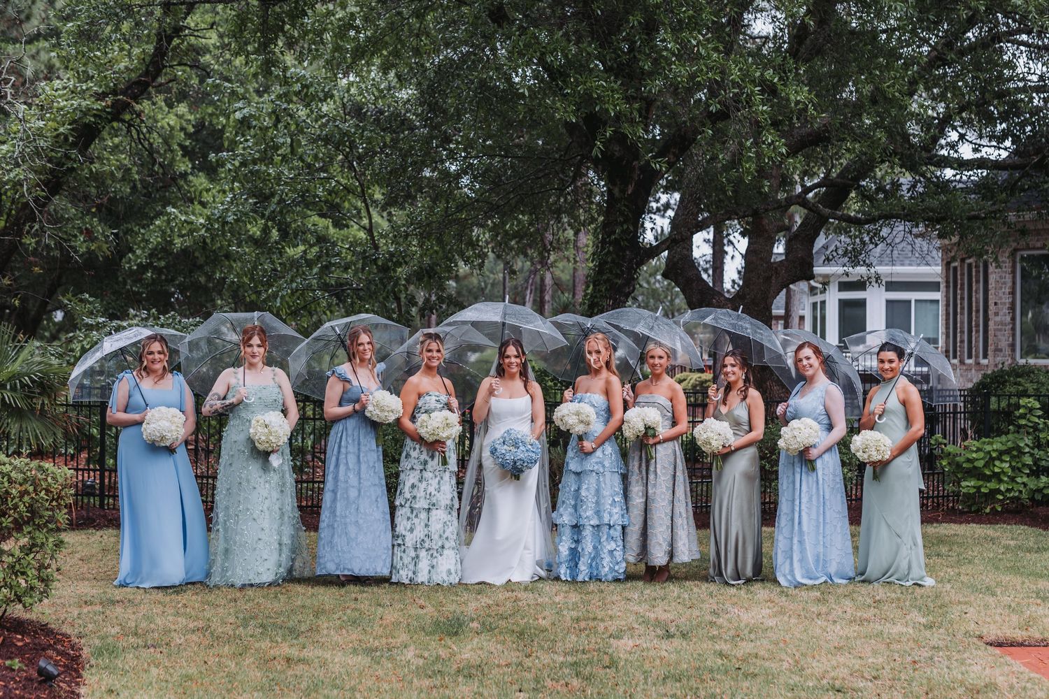 Bridal party in blue and green gowns holding clear umbrellas and white bouquets in garden.