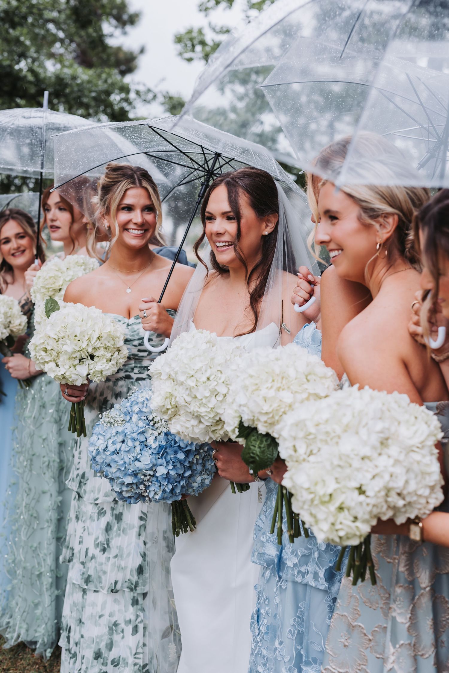Bride and bridesmaids in blue and sage dresses holding bouquets under clear umbrellas in rain.