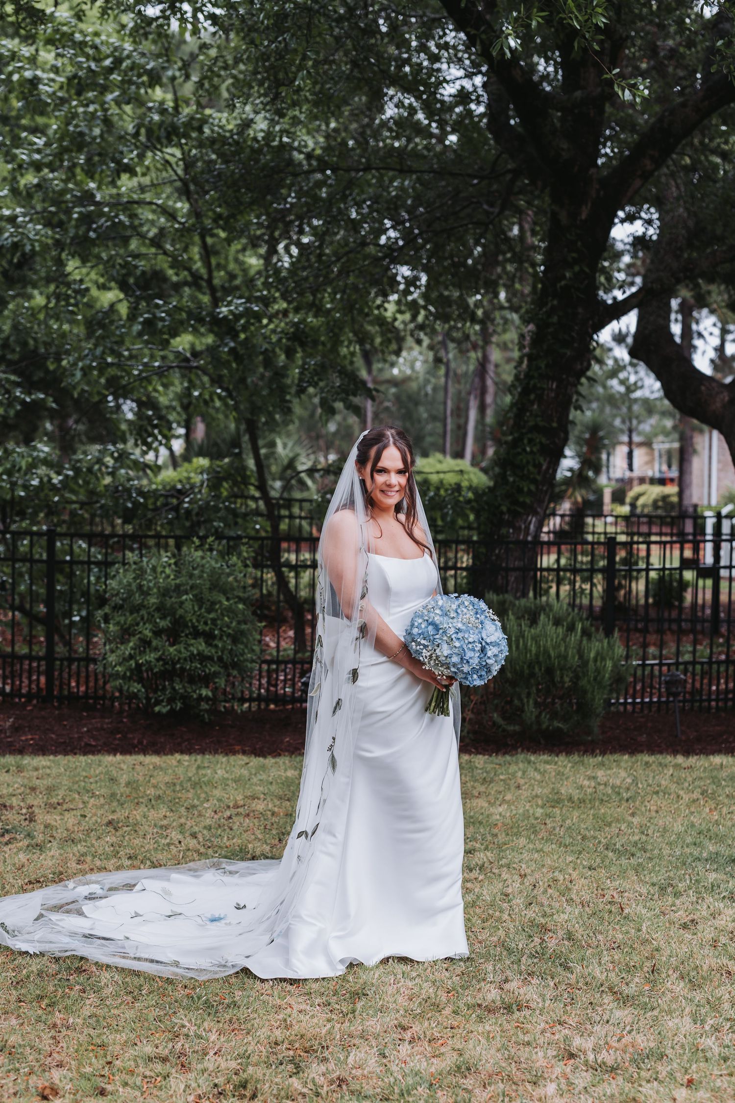 Bride in elegant white gown and cathedral veil holds blue hydrangea bouquet while posing in lush garden setting.