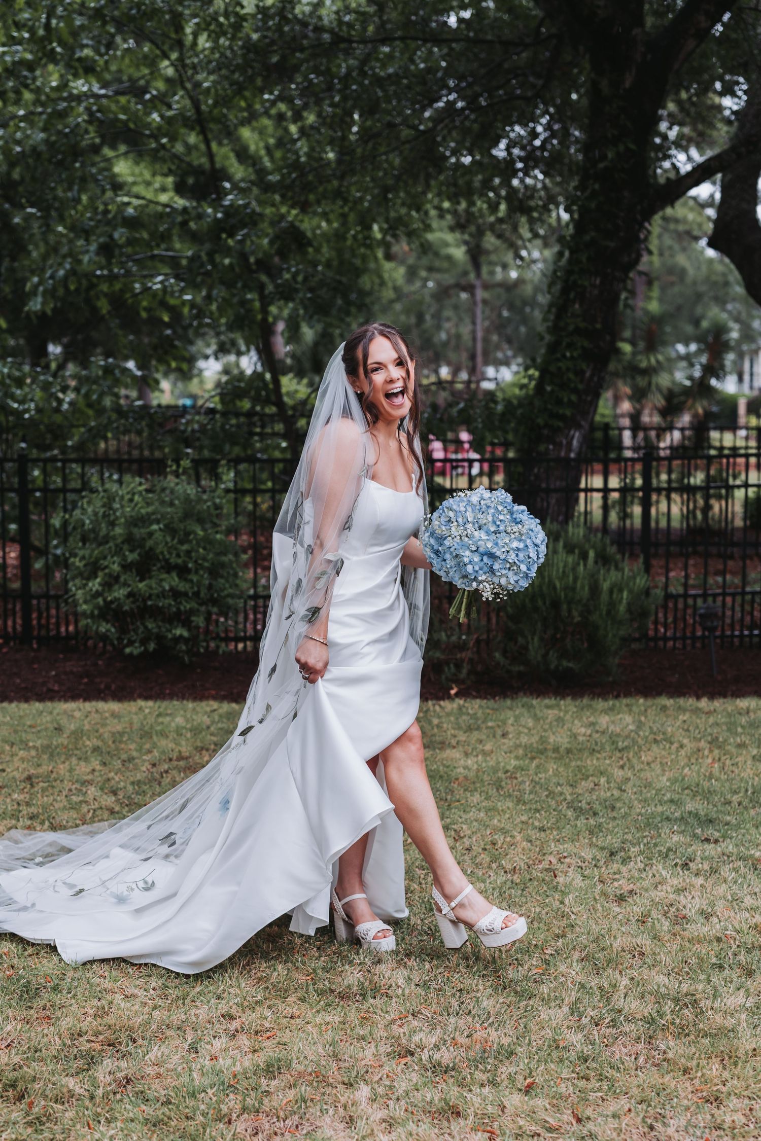Joyful bride in white gown with floral veil kicking up her heel on garden lawn.