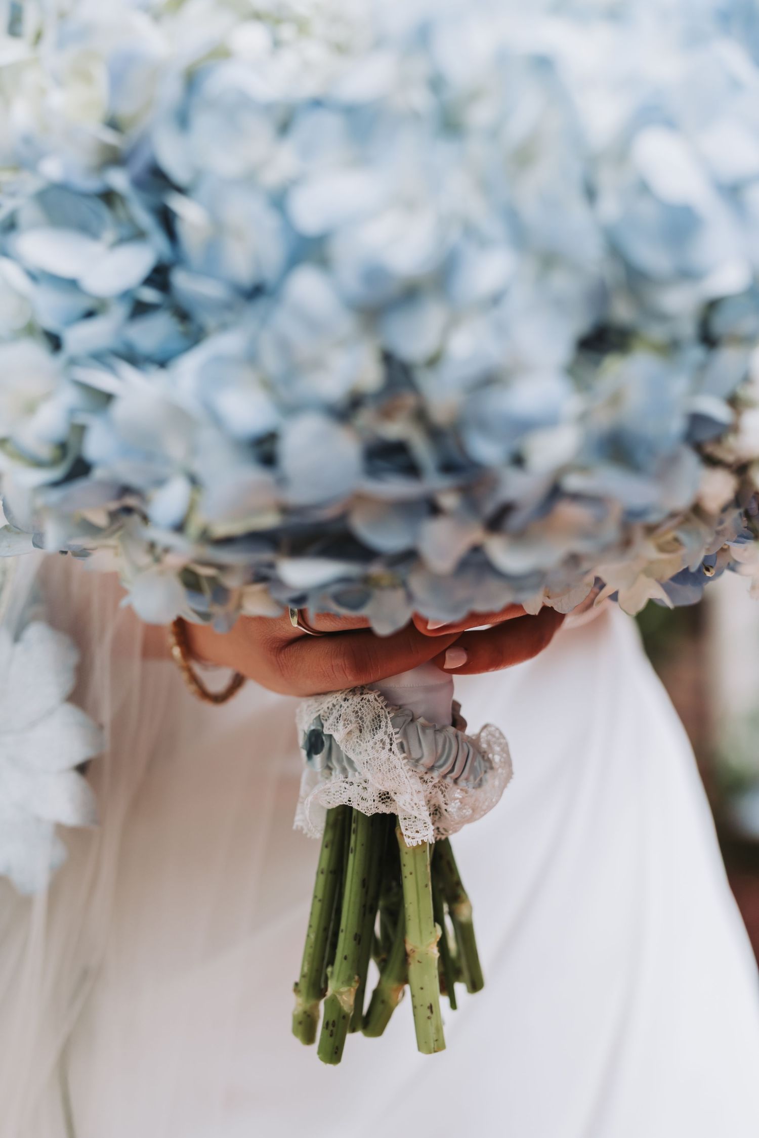 Close-up of lush blue hydrangea bridal bouquet wrapped in white lace ribbon held by bride in white dress.