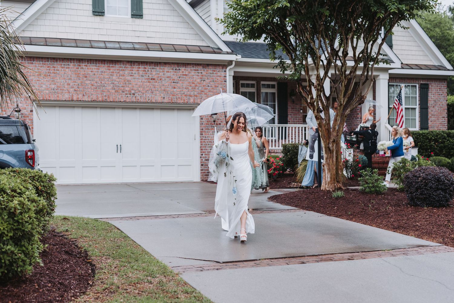 Bride in white jumpsuit walking down driveway holding a clear umbrella on a cloudy wedding day.