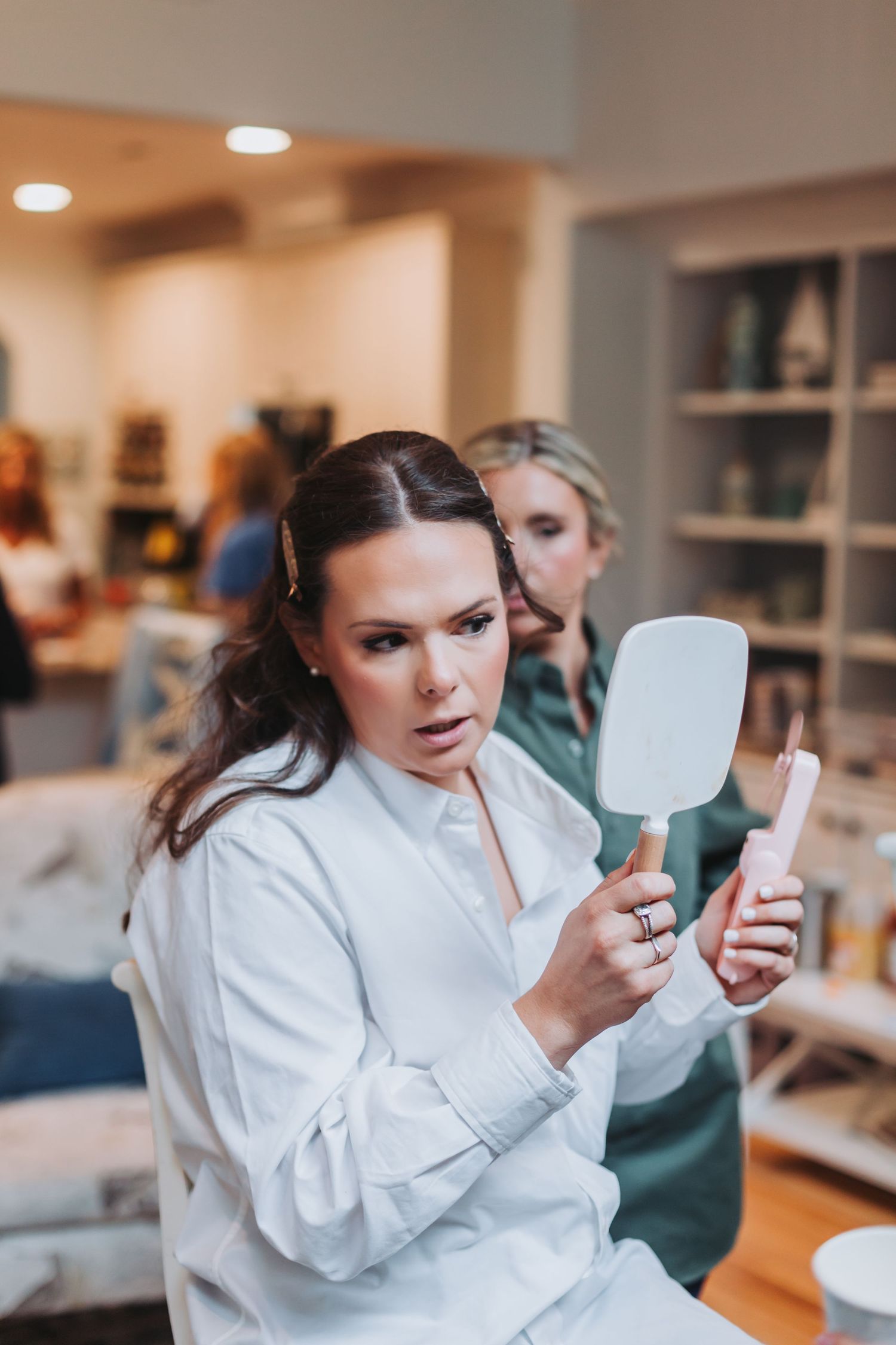 Bride in white robe examines makeup in handheld mirror while makeup artist works in bright salon setting.