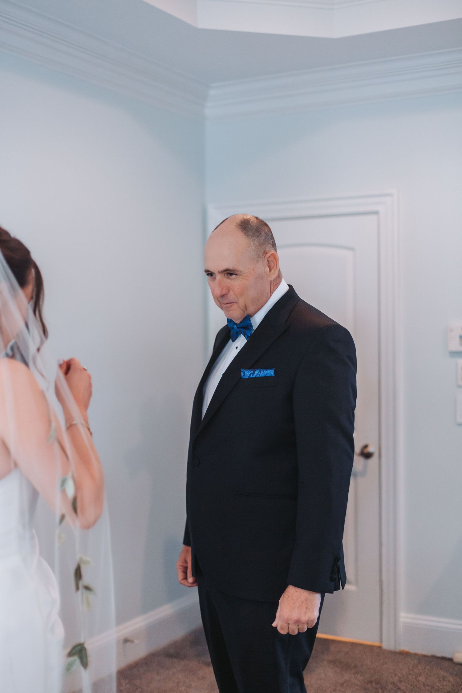 Father of the bride in navy tuxedo with blue bow tie shares emotional first look moment with bride in white gown.