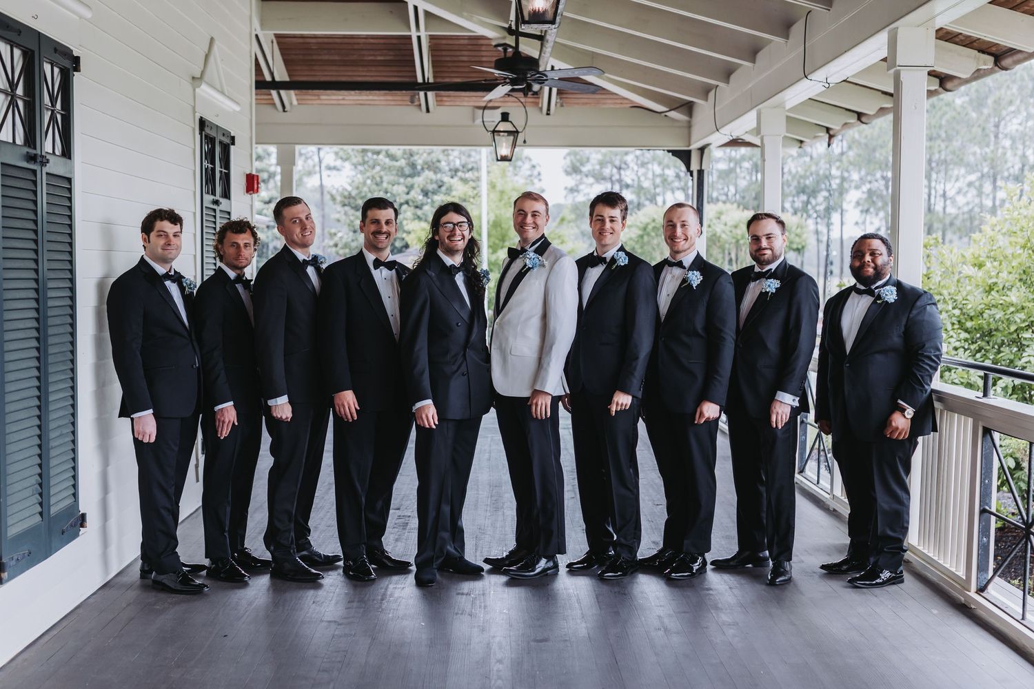 Groom in white jacket with groomsmen in black tuxedos posing on covered outdoor porch.